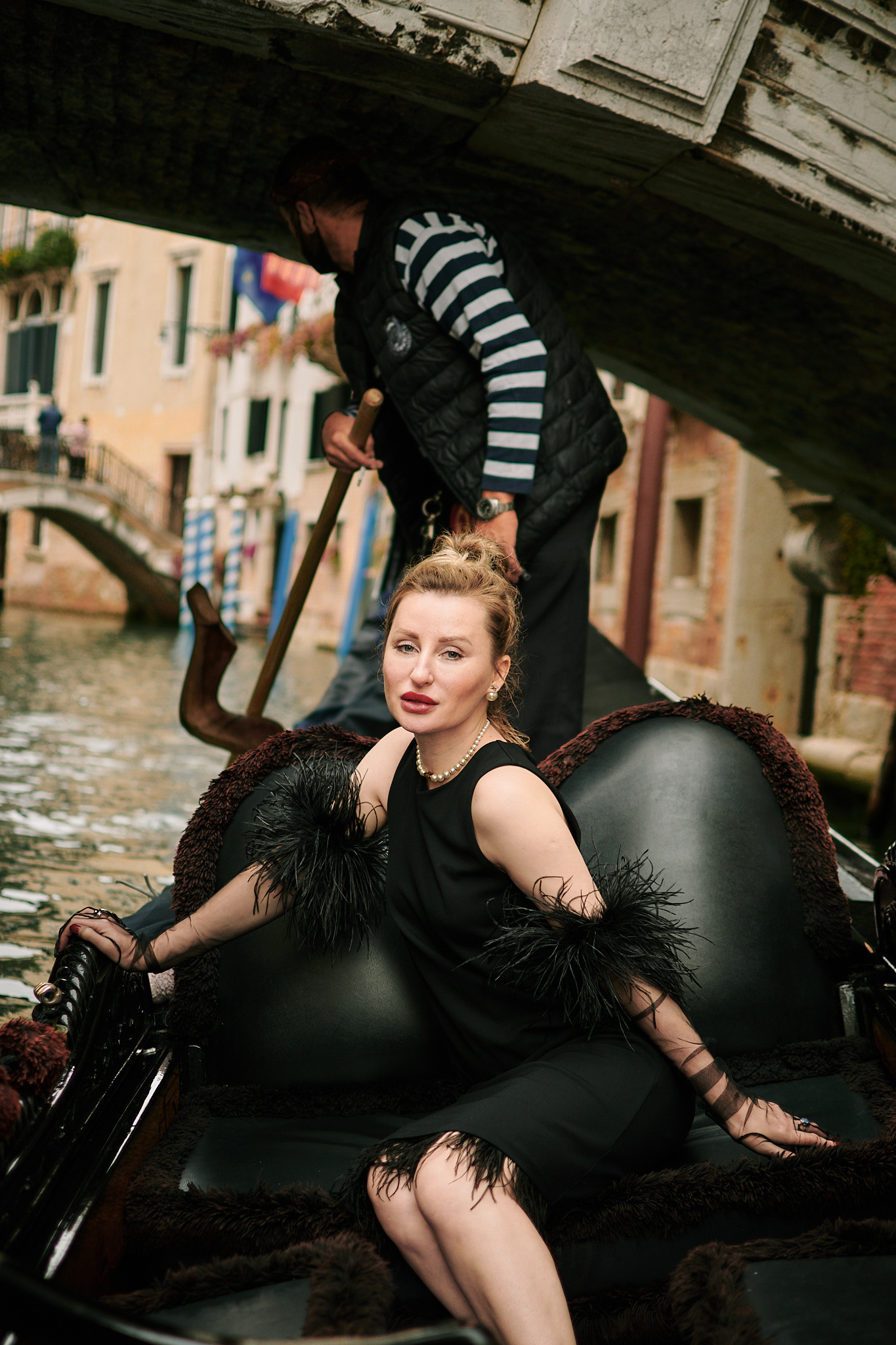 woman in black dress on gondola in Venice, Italy. Portrait of a young woman in a black dress standing on San Marco Square, Venice, with a breathtaking panoramic view of the Laguna in the background. The woman has long brown hair styled in loose waves, and she is looking dreamily towards the view. The square is bustling with people, and the stunning architecture of the surrounding buildings adds to the romantic atmosphere of the location. Fashion blogger shoot Venice. Portrait and Fashion Photographer in Venice, Italy. Instagram Photos with lifestyle photographer