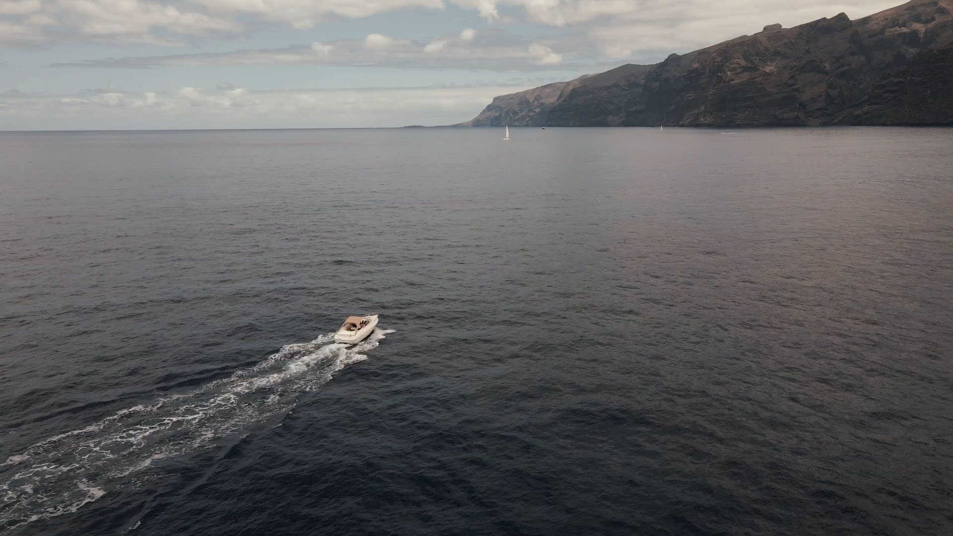 A small boat sailing across a calm sea under a partly cloudy sky, with a mountainous coastline of Los Gigantes, Tenerife in the distance.