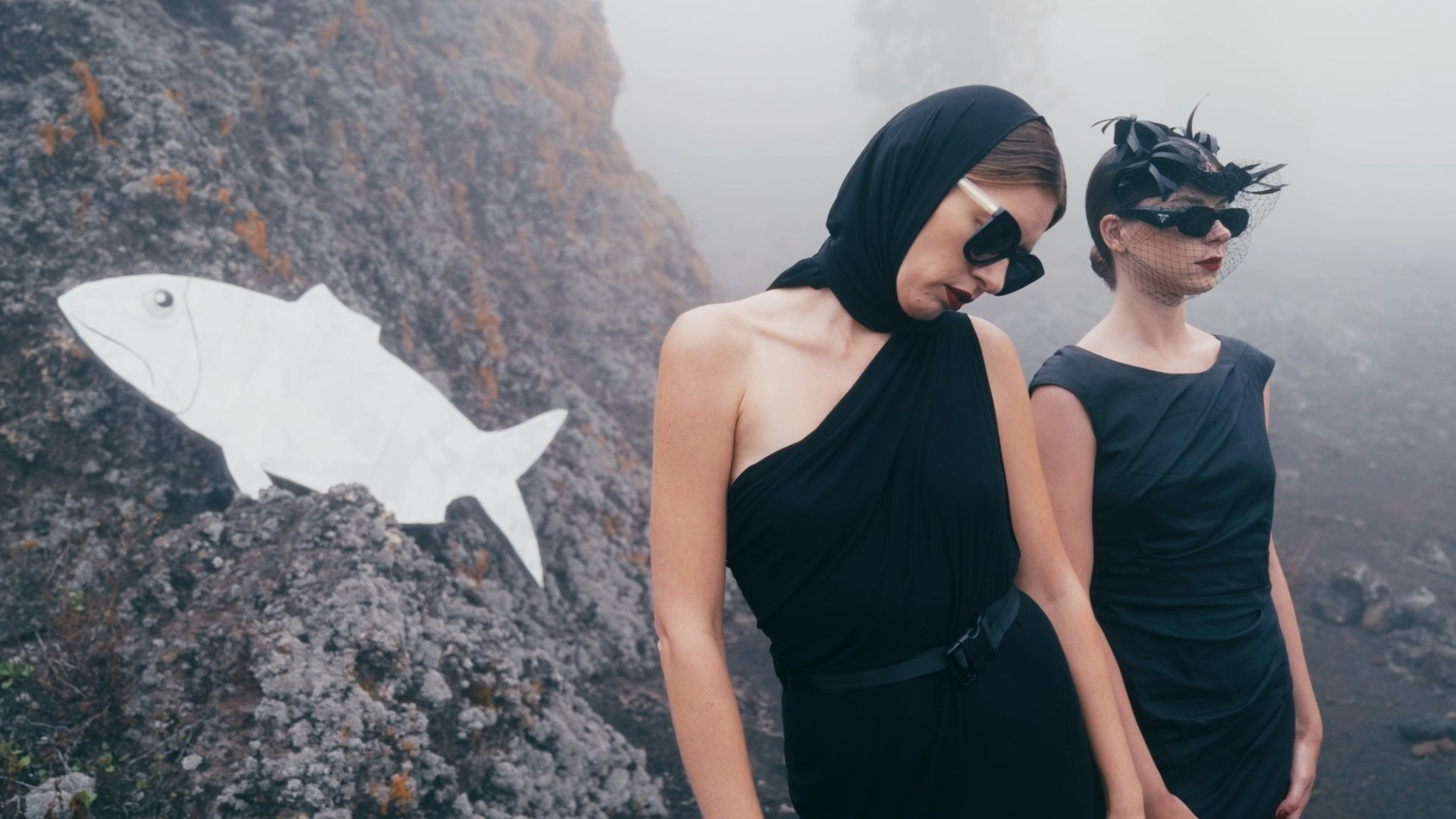 Two women dressed in avant-garde black outfits pose against volcanic rocks. A white paper fish lies on the ground beside them, adding a surreal and conceptual element.