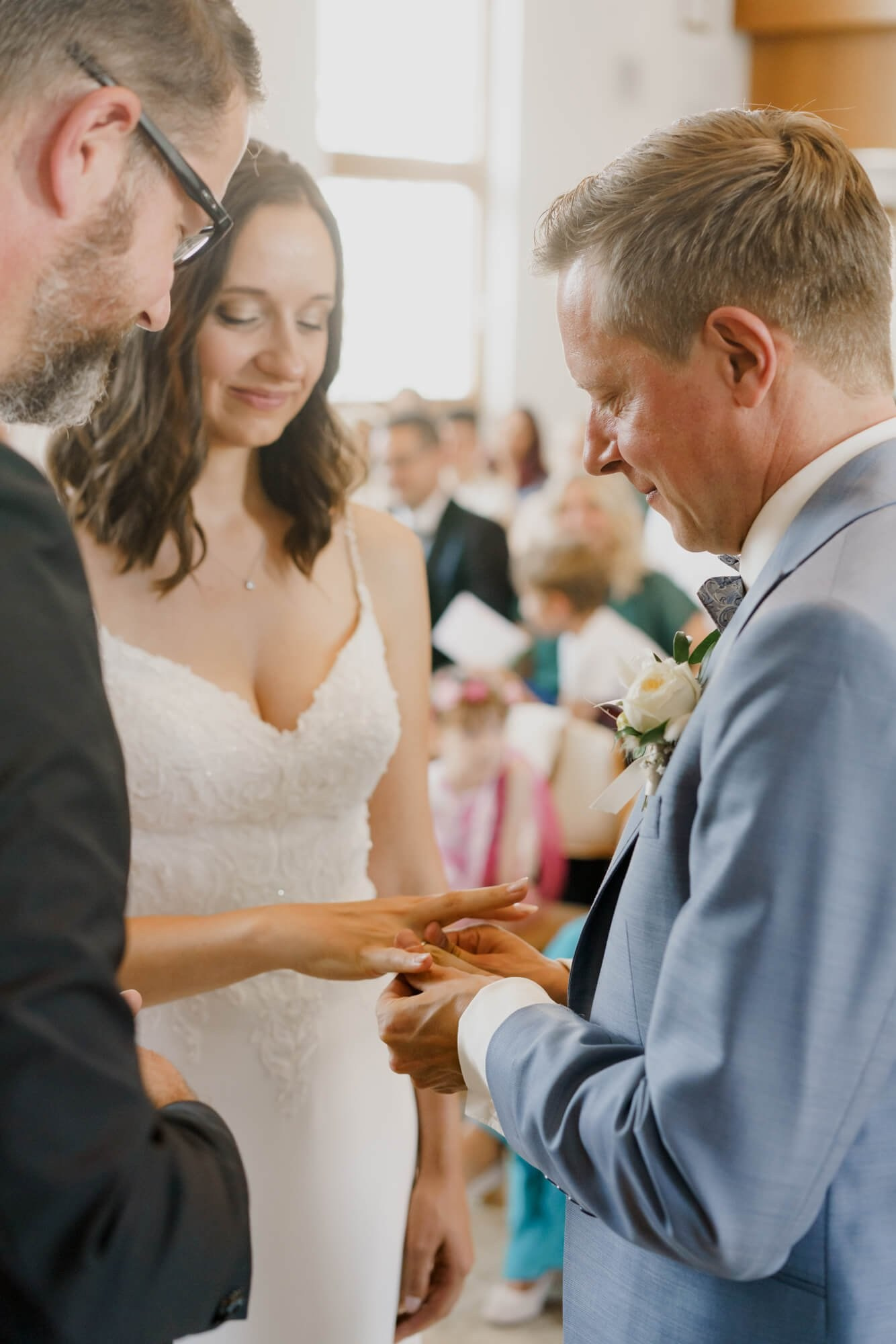 Groom sliding a wedding ring onto the bride's finger during the ring exchange, guests softly blurred behind them