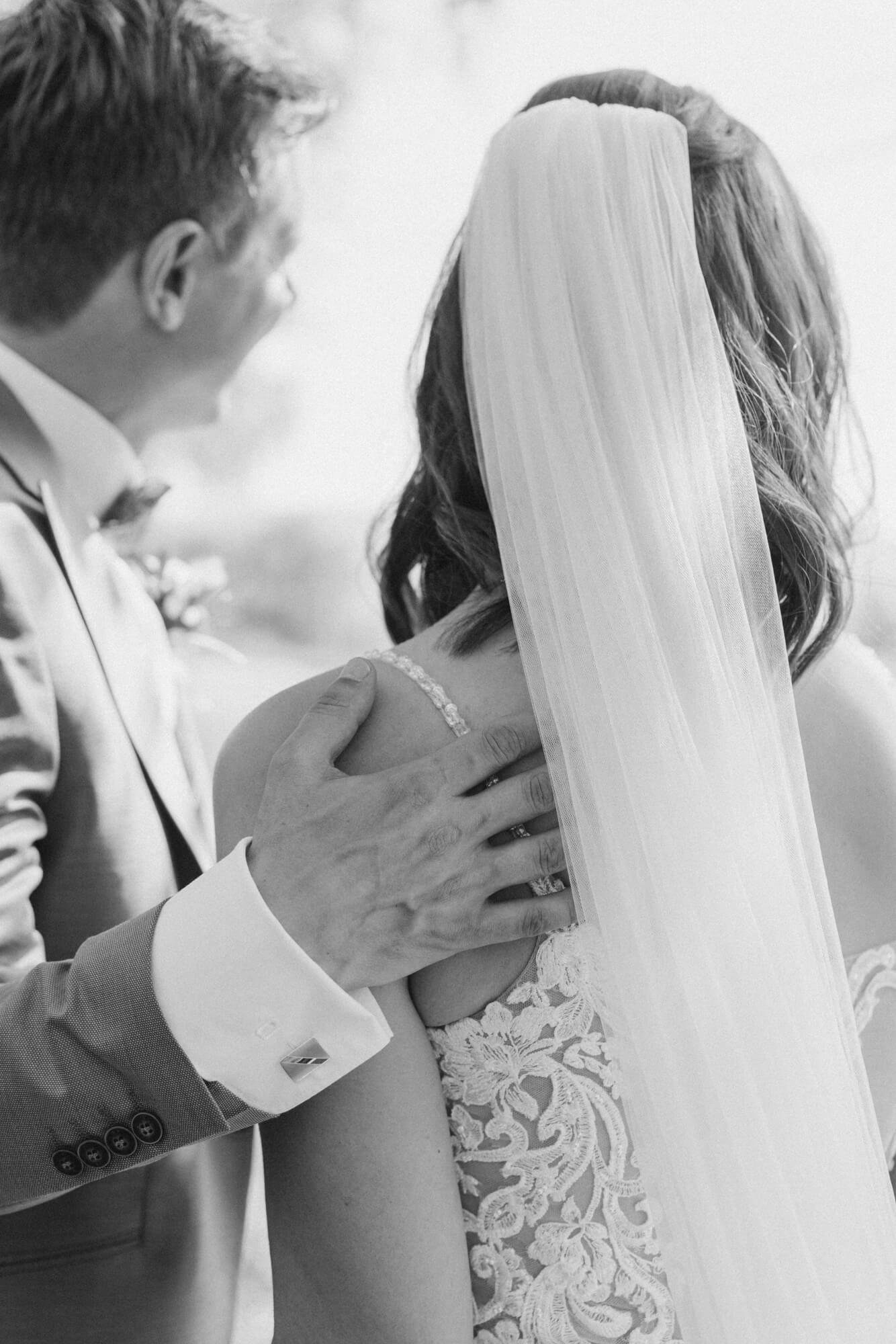 Groom embracing the bride from behind, her lace bodice and cathedral veil in close focus; black and white
