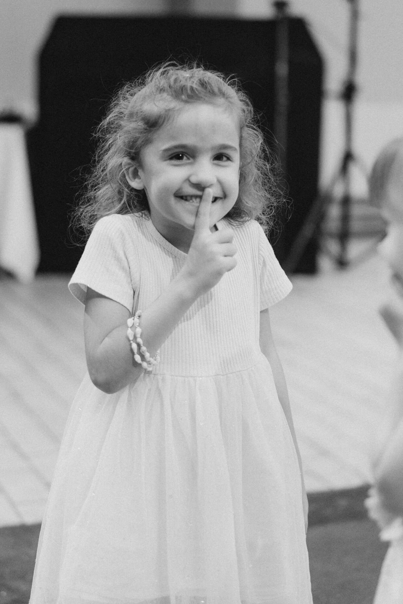 A young girl in a white dress with curly hair pressing a finger to her lips during the wedding reception