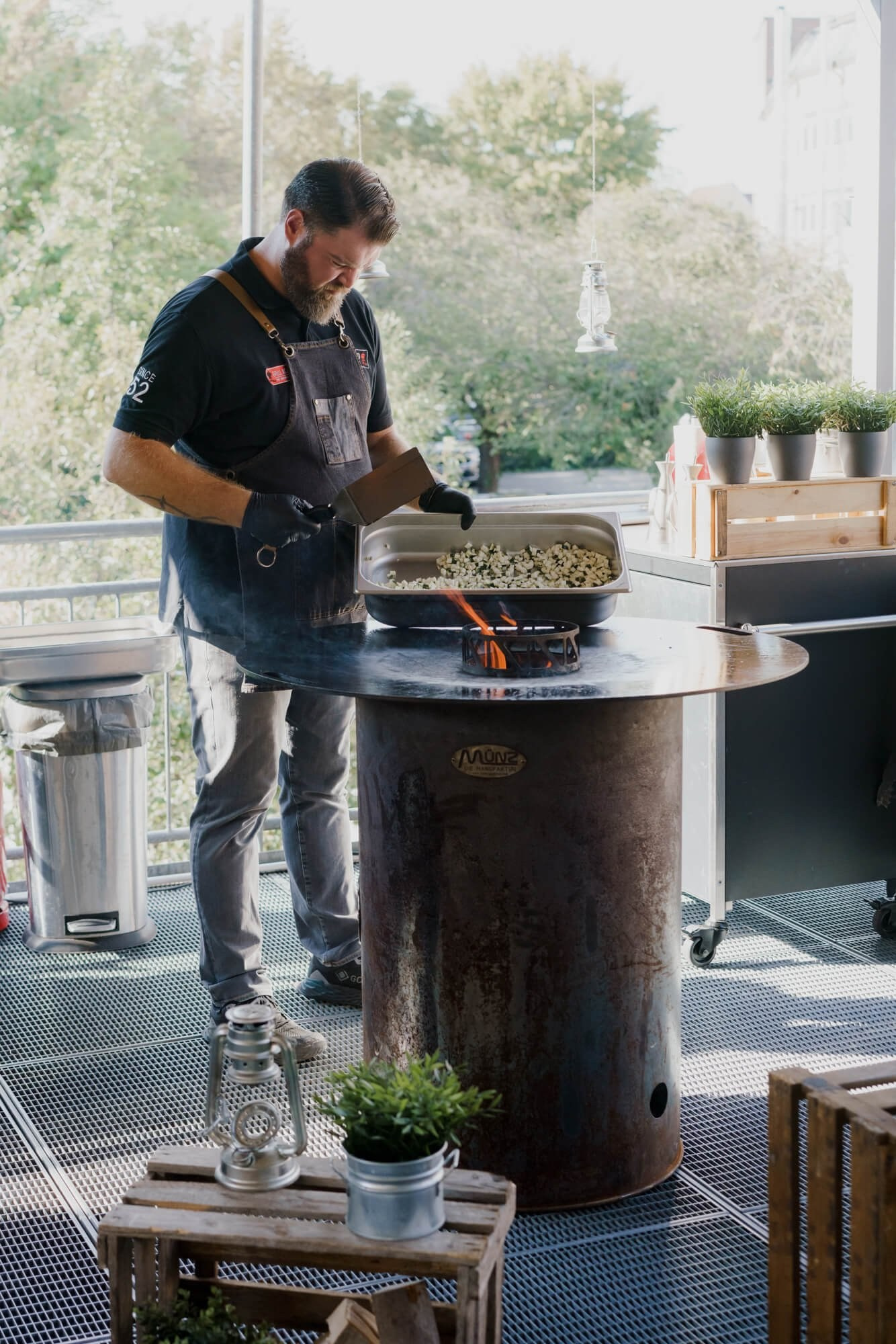 A caterer tending a large outdoor grill on the reception venue terrace, trees visible through floor-to-ceiling windows behind