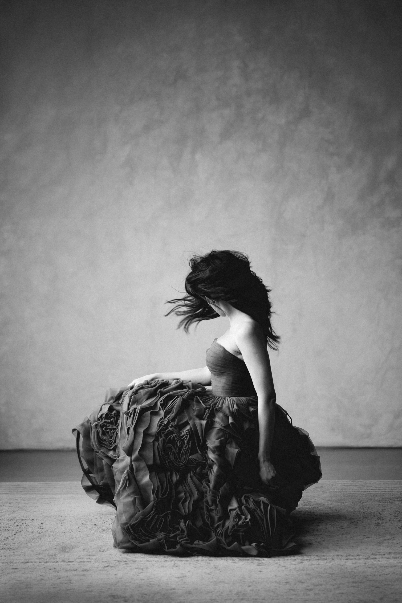 crno bela fotografija mlade žena u prekrasnoj haljini na karenere čuči i uhvaćena u trenutku okreta glava, gde joj kosa prelazi preko čelog lica