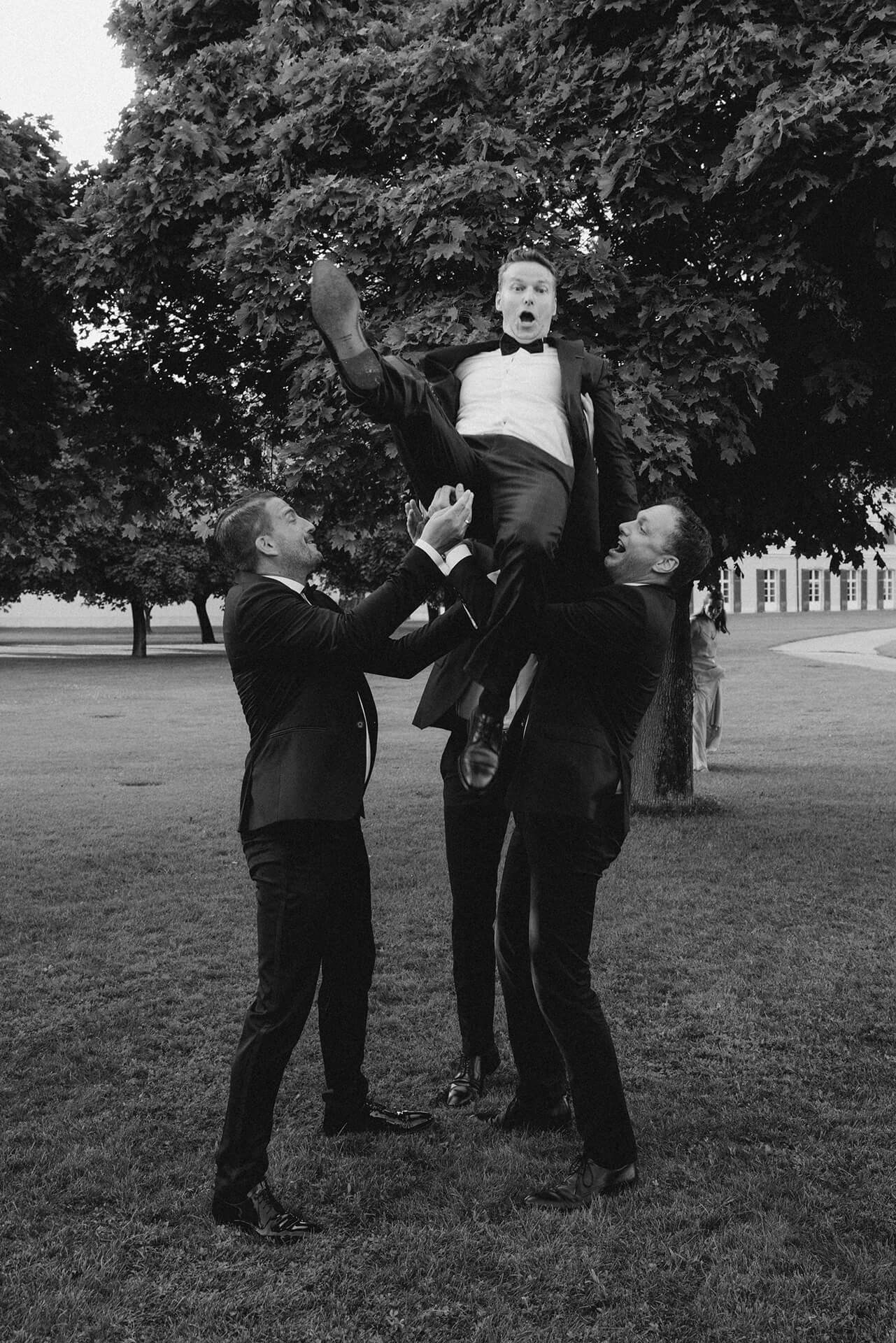 Groomsmen toss groom in air on lawn, black tie wedding