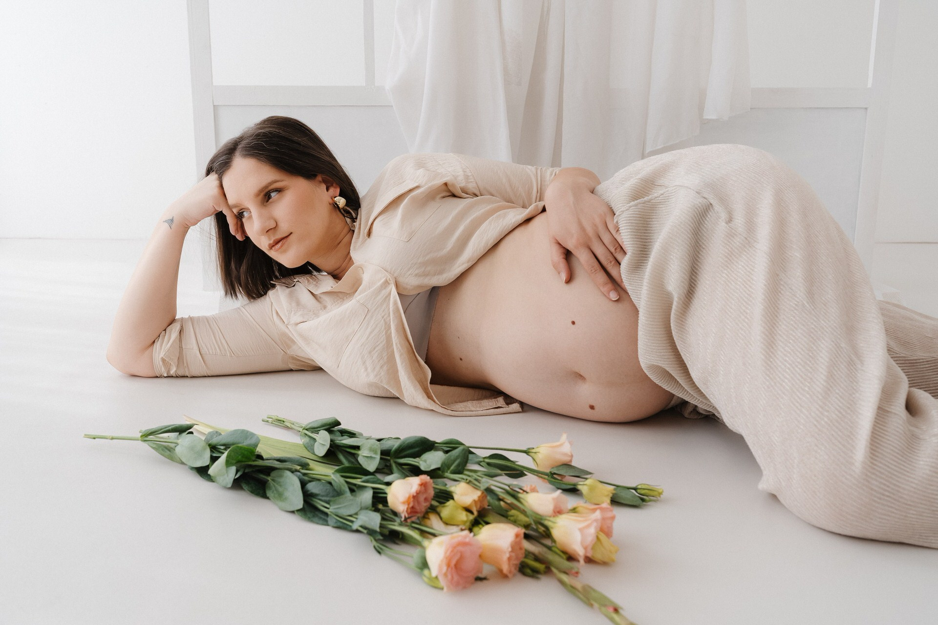 Trudnica leži na boku u svetlom enterijeru, oslonjena na ruku, dok nežno drži stomak, a ruže su položene ispred nje na podu.
