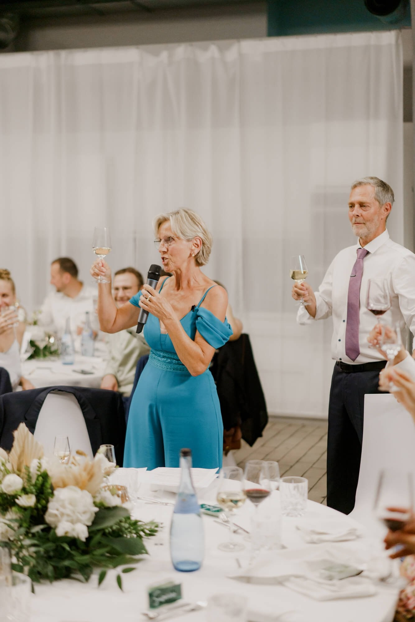 A guest in a teal dress giving a toast with a microphone while another guest raises a wine glass behind her, floral centerpieces on the tables