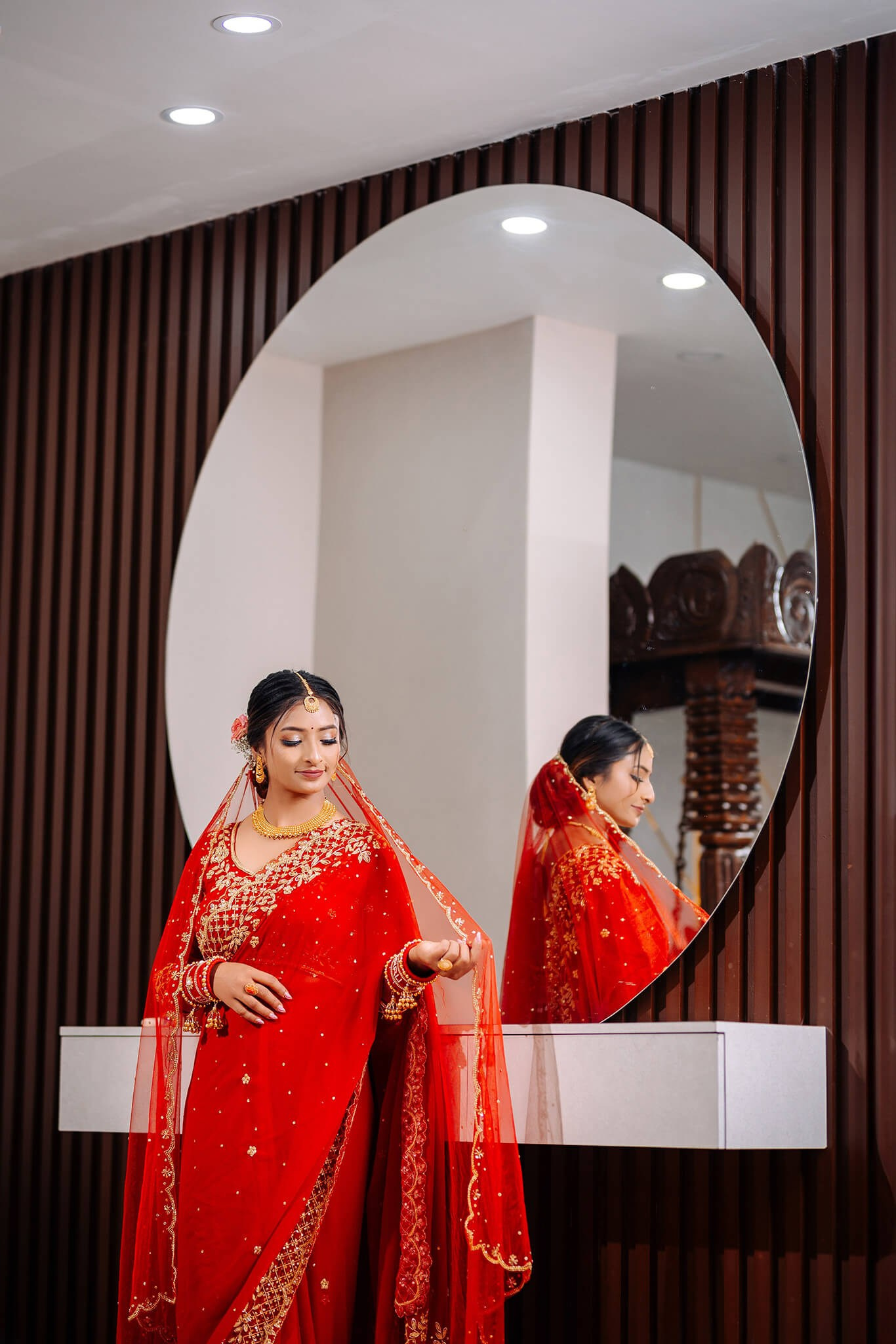 Beautiful Nepali bride in a traditional red wedding dress looking at her reflection in a circular mirror.