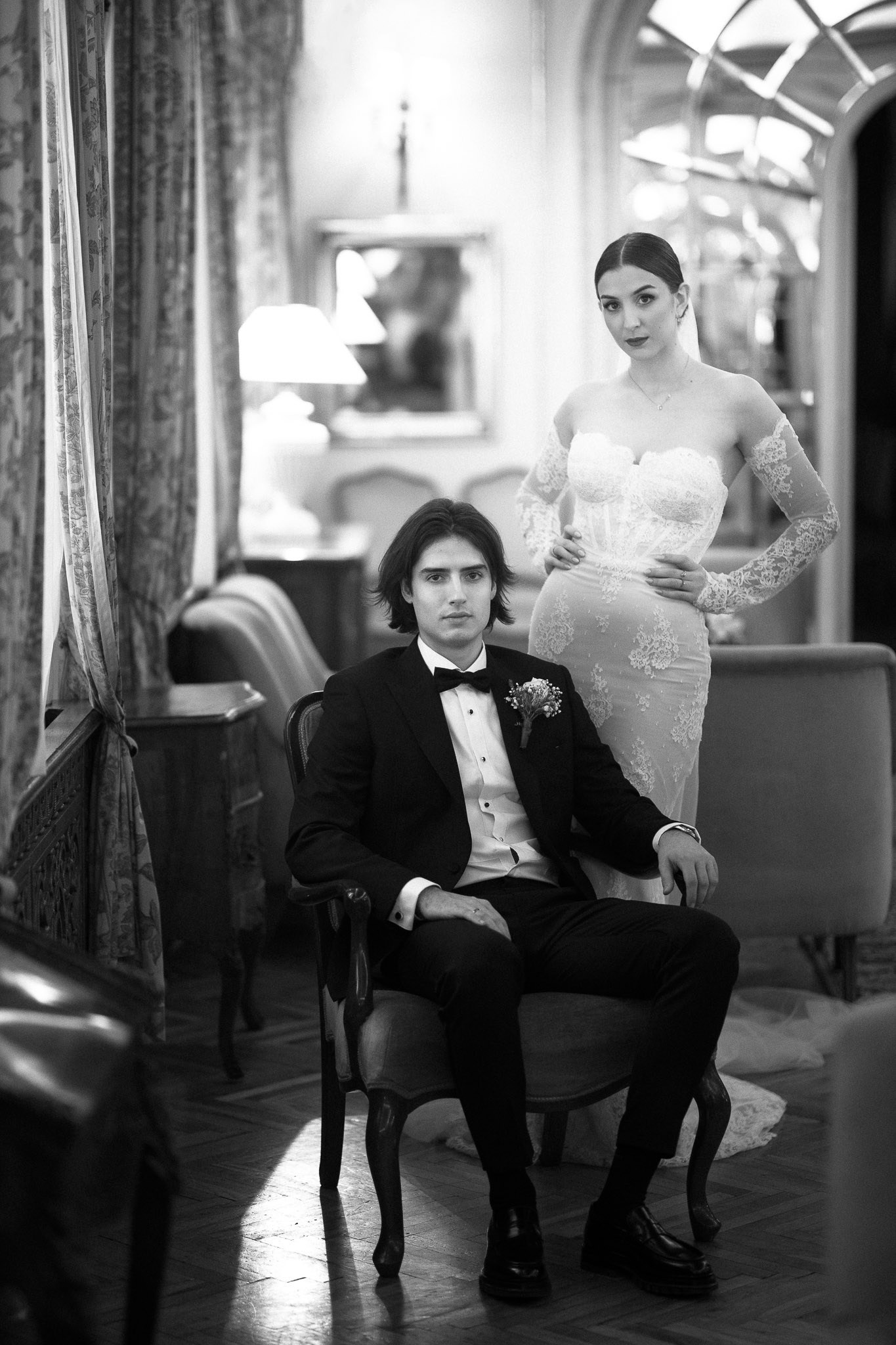 Crno-bela fotografija venčanja: mlada u čipkanoj venčanici sa odvojenim rukavima stoji iza mladoženje u tuksidu koji sedi. Editorijal u Aeroklubu