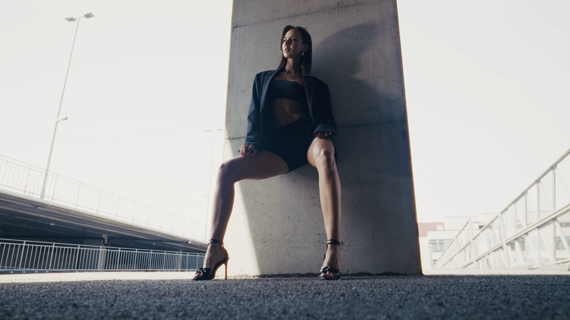 A person sitting against a concrete column outdoors, wearing a dark jacket and sportswear, with a confident and relaxed posture.
