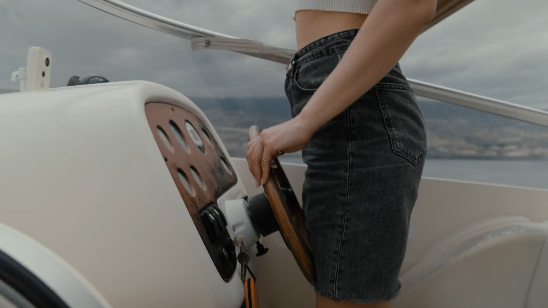  A close-up view of a person standing at the helm of a boat, their hand resting on the steering wheel, wearing a casual outfit of a crop top and denim shorts, with the sea and Tenerife coastline visible in the background.
