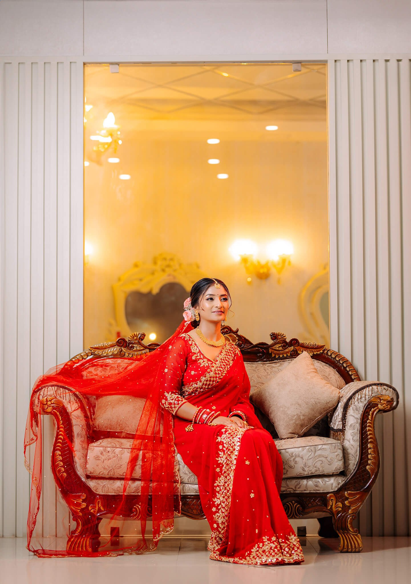 A graceful Nepali bride dressed in a traditional red sari sitting elegantly on a vintage-style sofa.