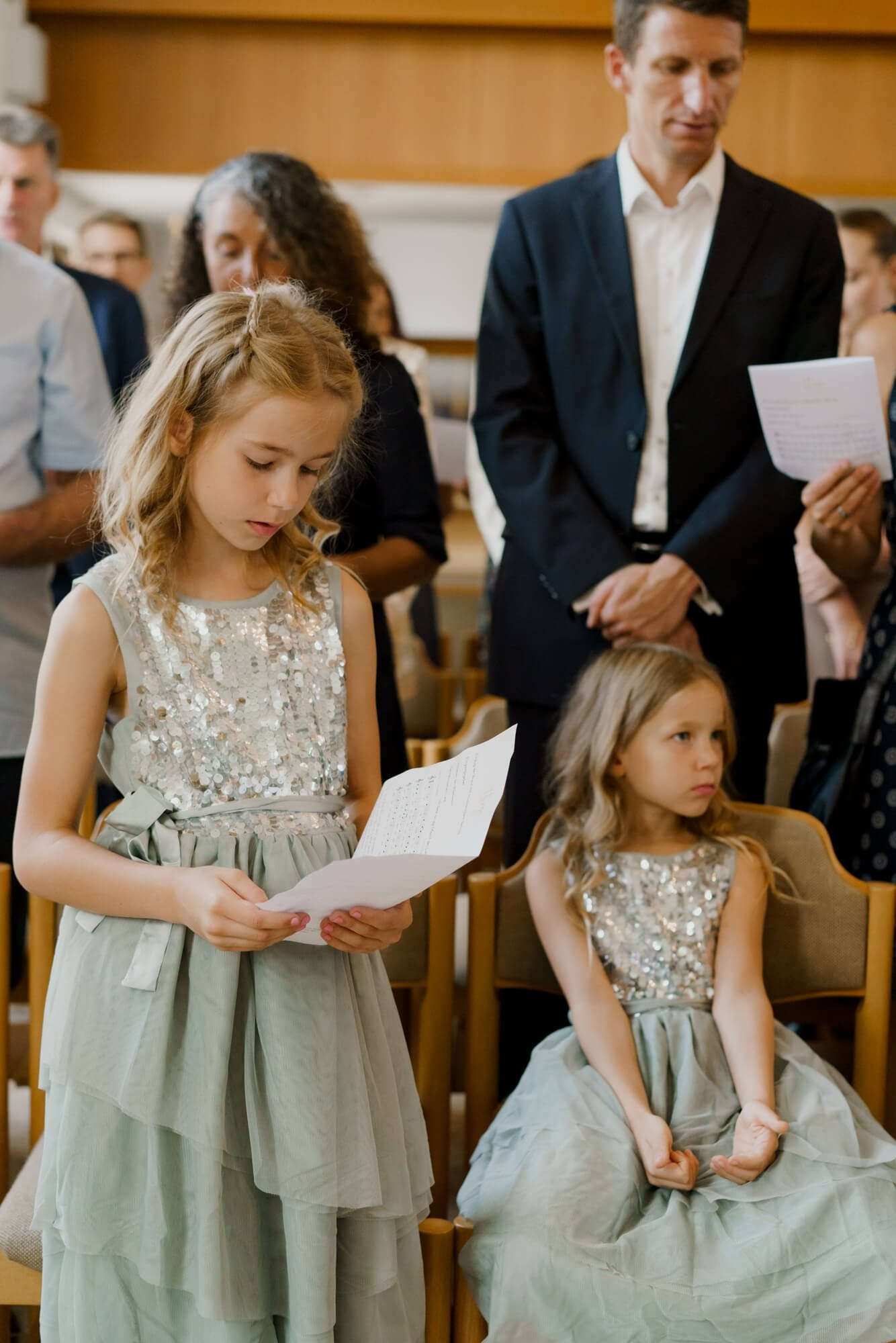 Two young girls in sage green dresses seated in the ceremony hall, one holding a paper program and watching the service
