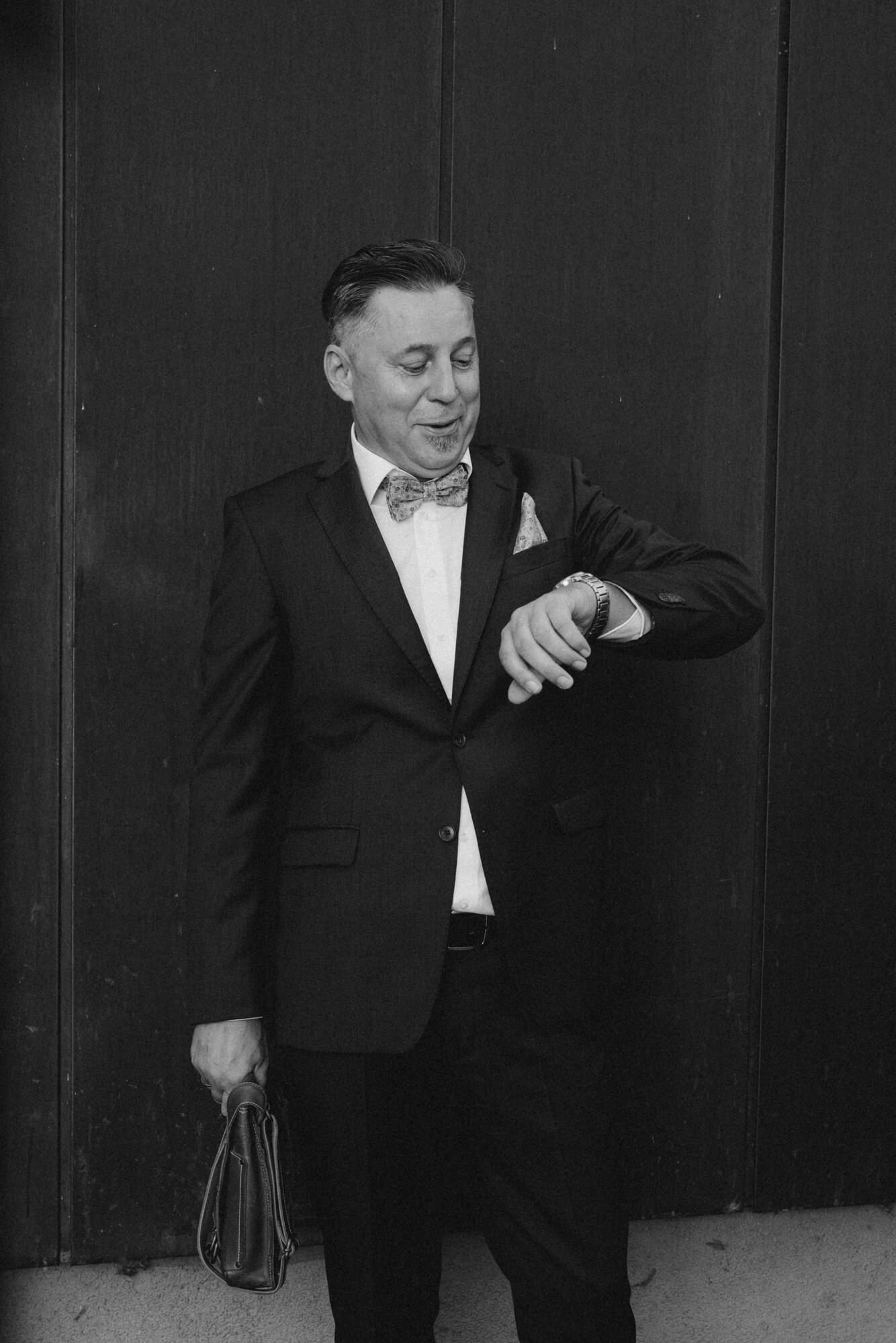A wedding guest in a bow tie checking his watch outside against a dark wall; black and white
