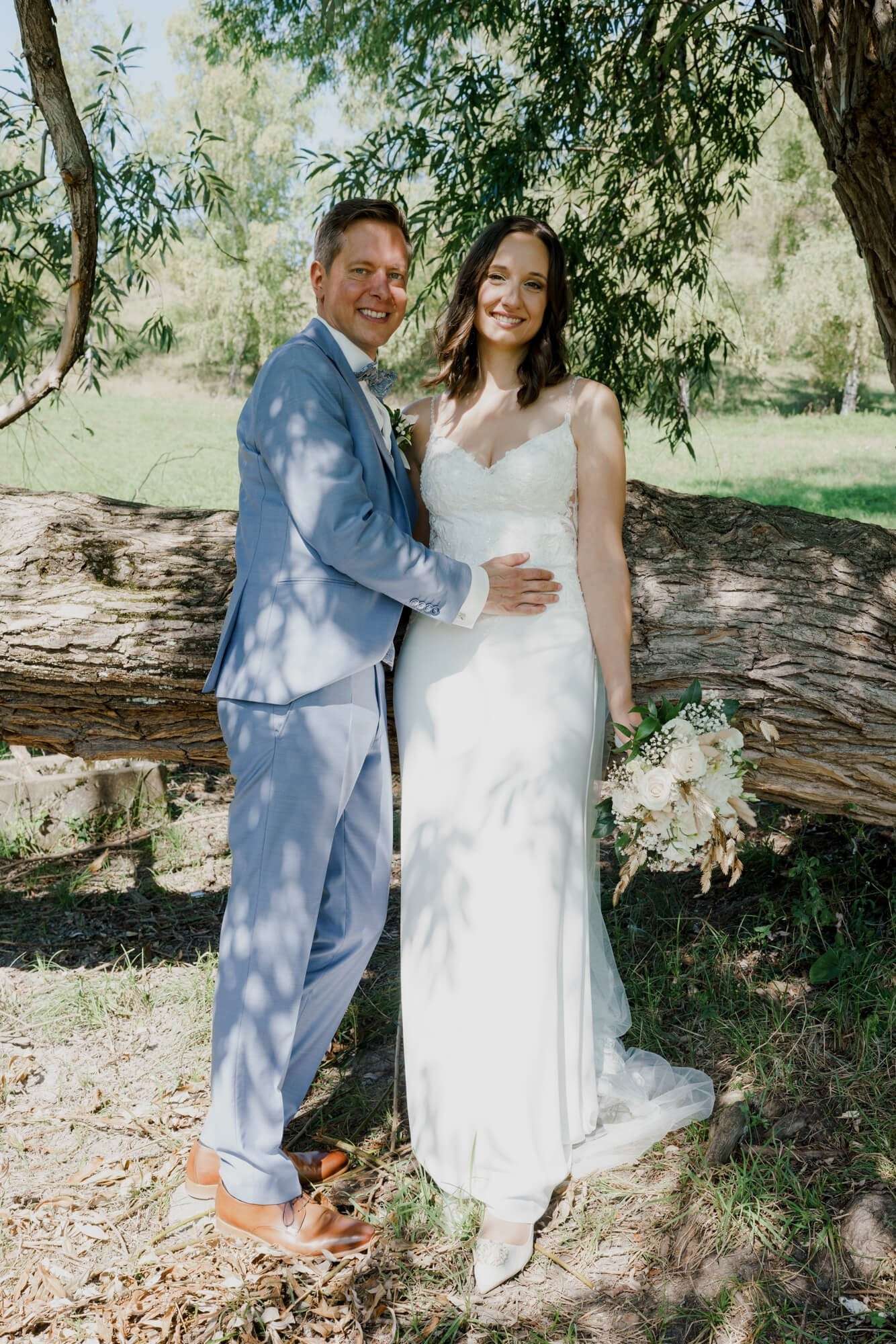 Couple posing together under a willow tree beside a large weathered log, bride holding a white bouquet