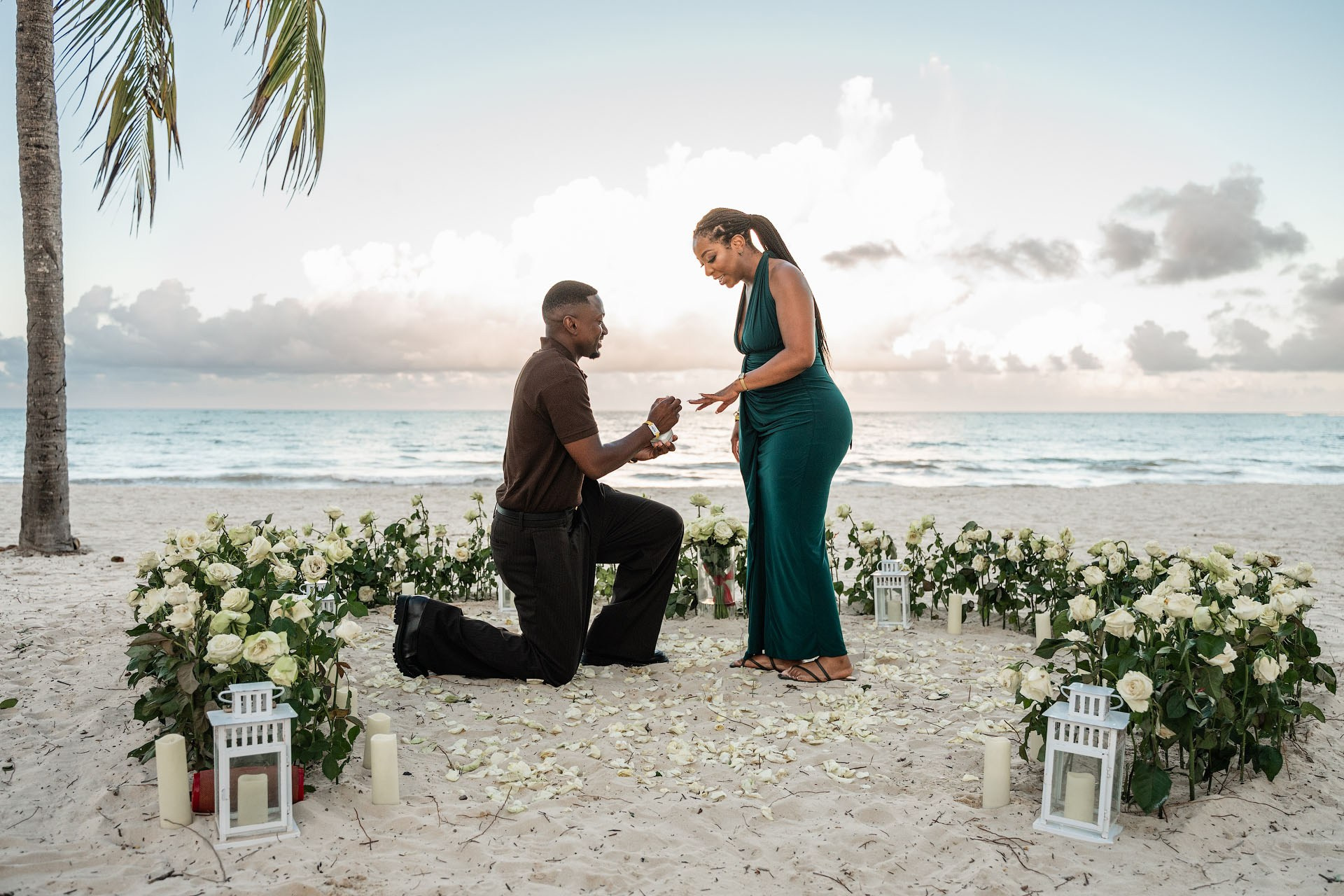 Man proposing on one knee to his smiling partner inside a large heart made of fresh white roses by the ocean.