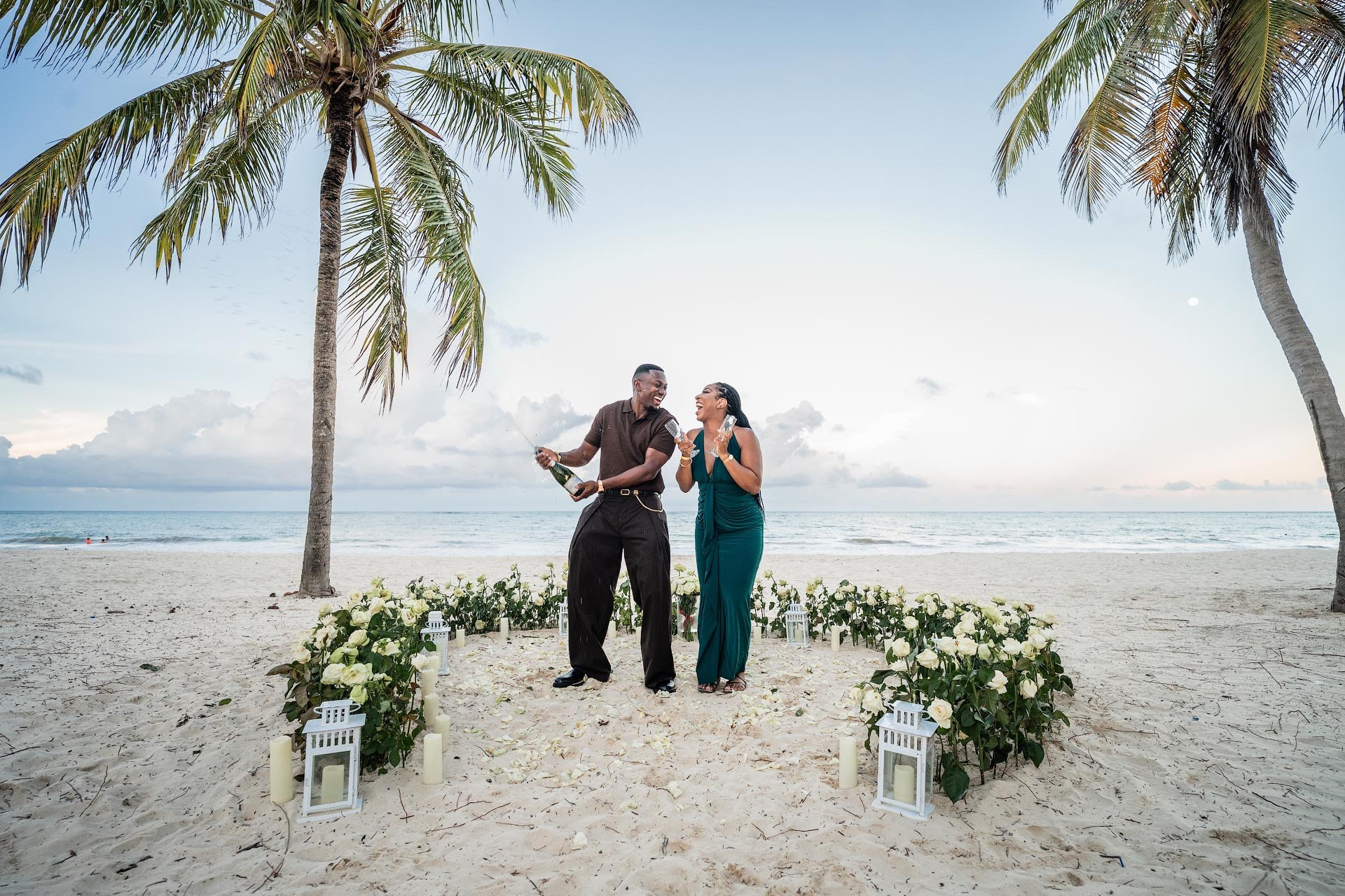 Couple laughing while spraying champagne beside their white rose heart proposal setup in Punta Cana.