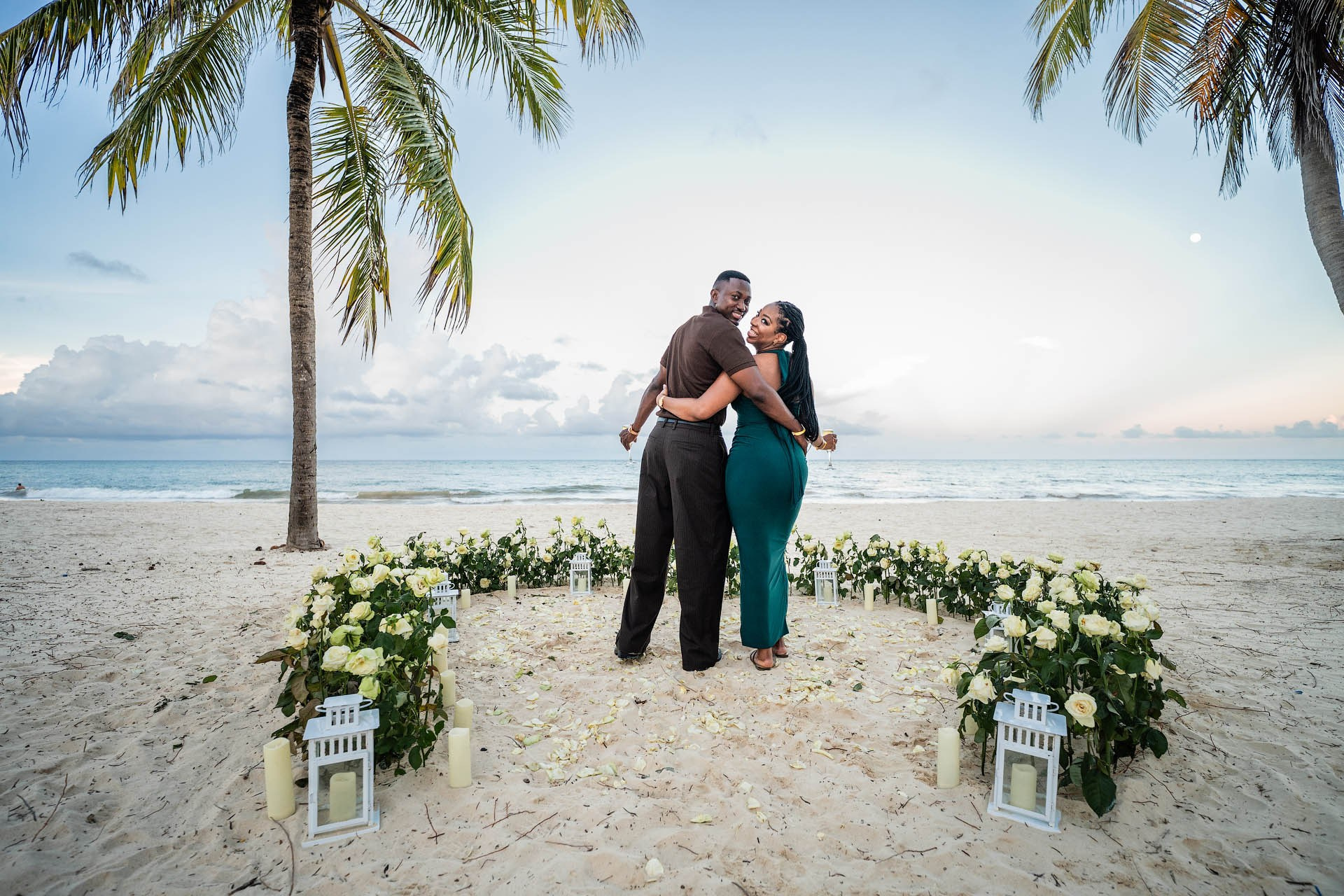 Engaged couple embracing inside a large heart of white roses beneath palm trees at Arena Gorda Beach.