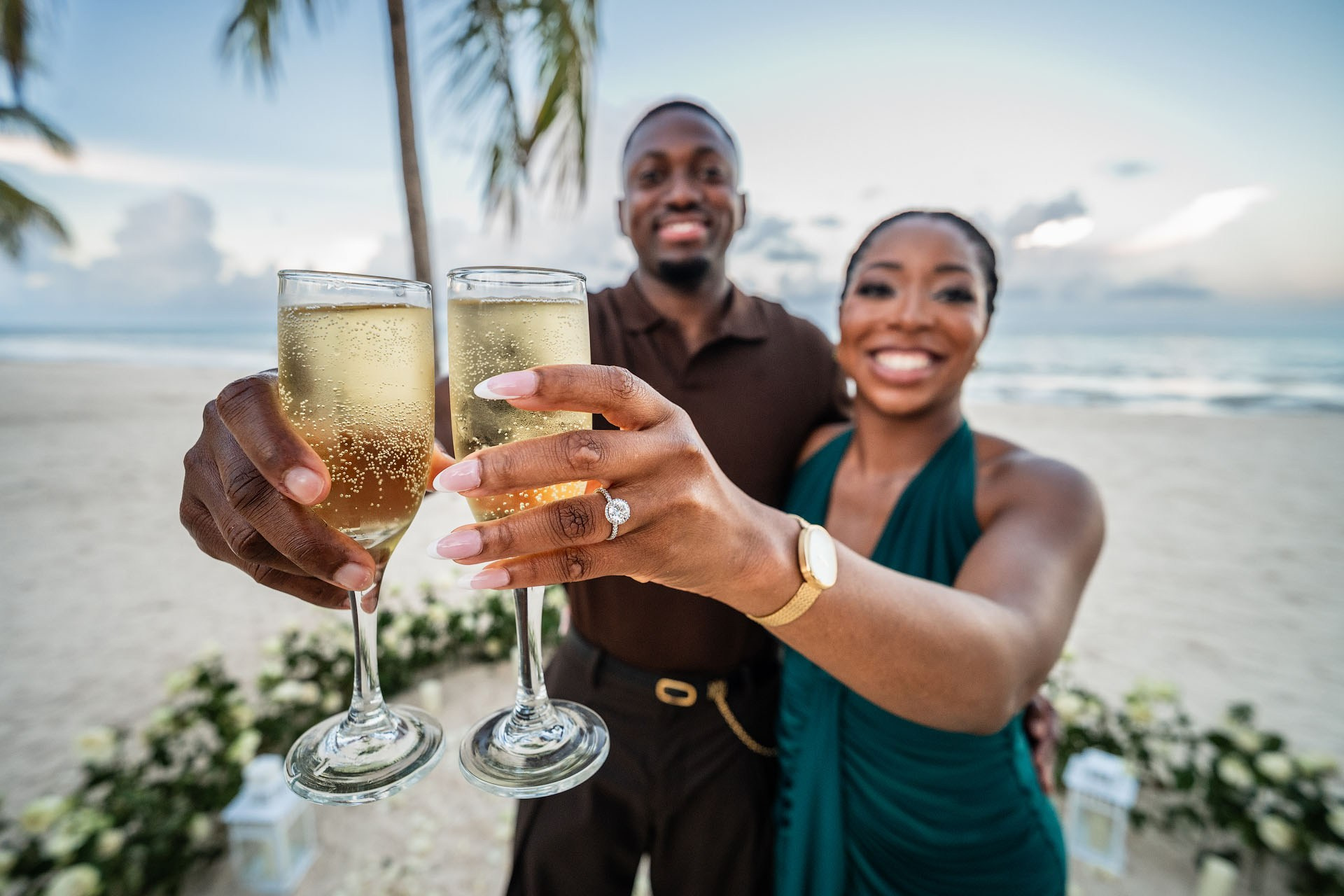Close-up of an engaged couple raising two champagne glasses with an engagement ring visible.