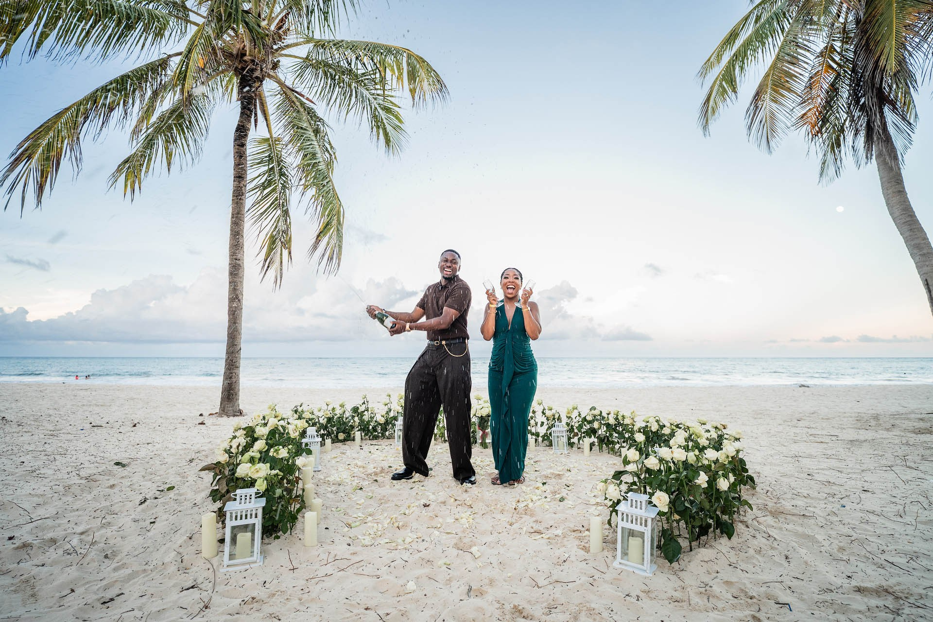 Joyful couple celebrating their engagement with champagne surrounded by fresh white roses and lanterns.