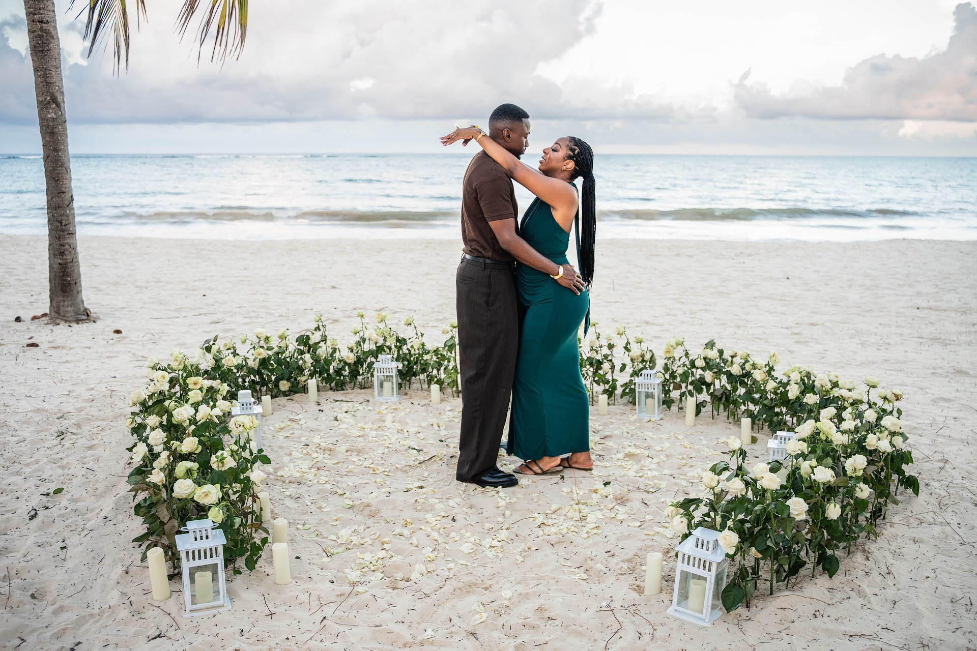 Engaged couple sharing a kiss inside a heart of fresh white roses on the sand.