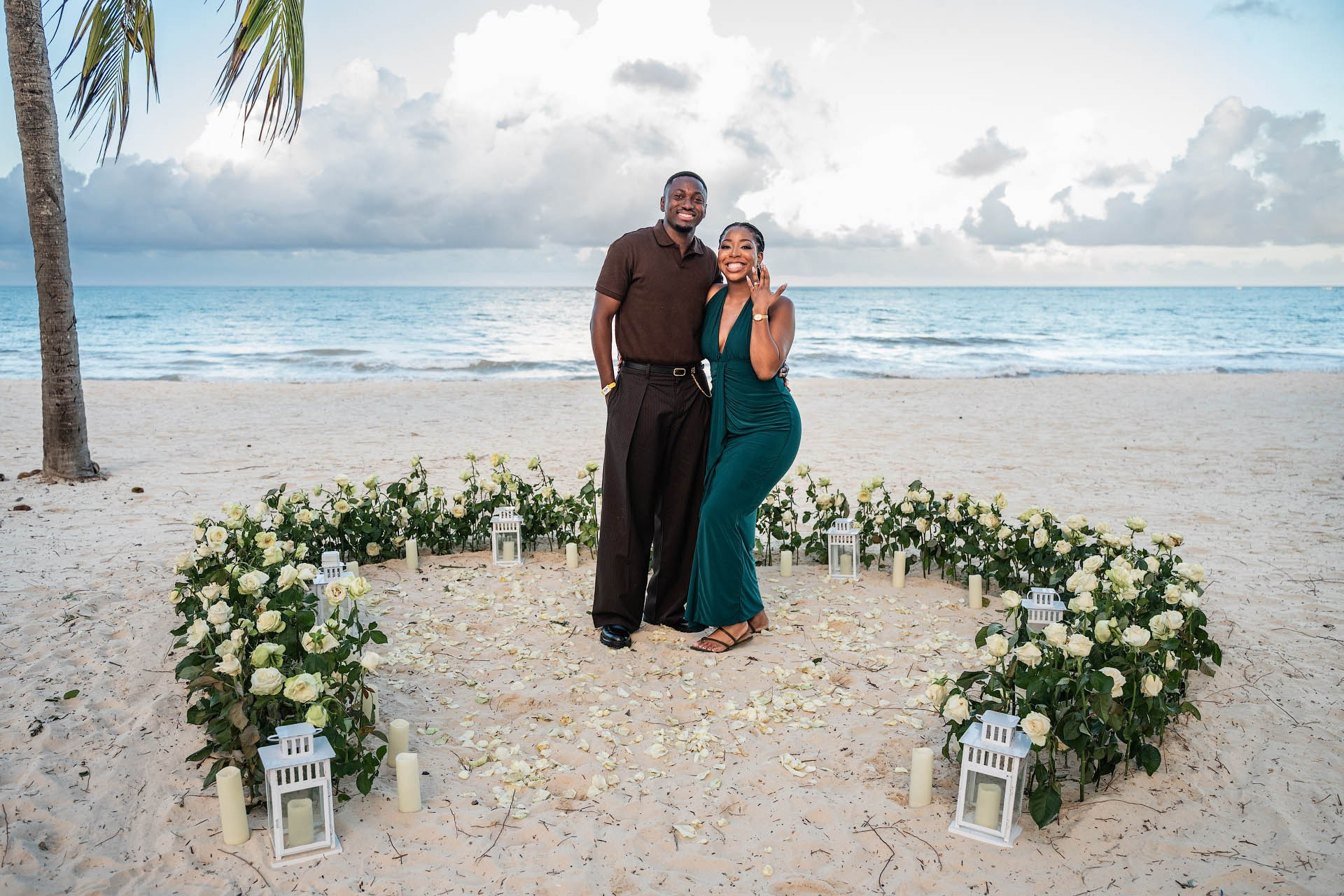 Happy couple posing among white roses and decorative lanterns after their beach marriage proposal.