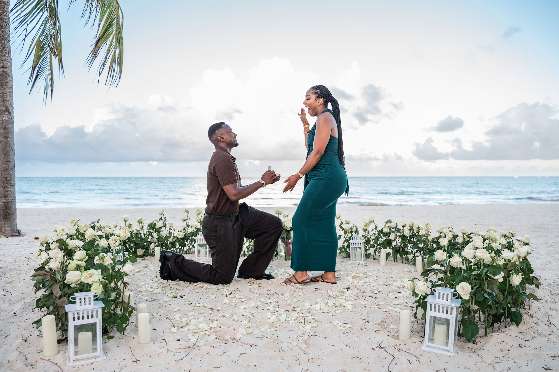 Man kneeling with an engagement ring while his surprised partner stands inside a heart of white roses.