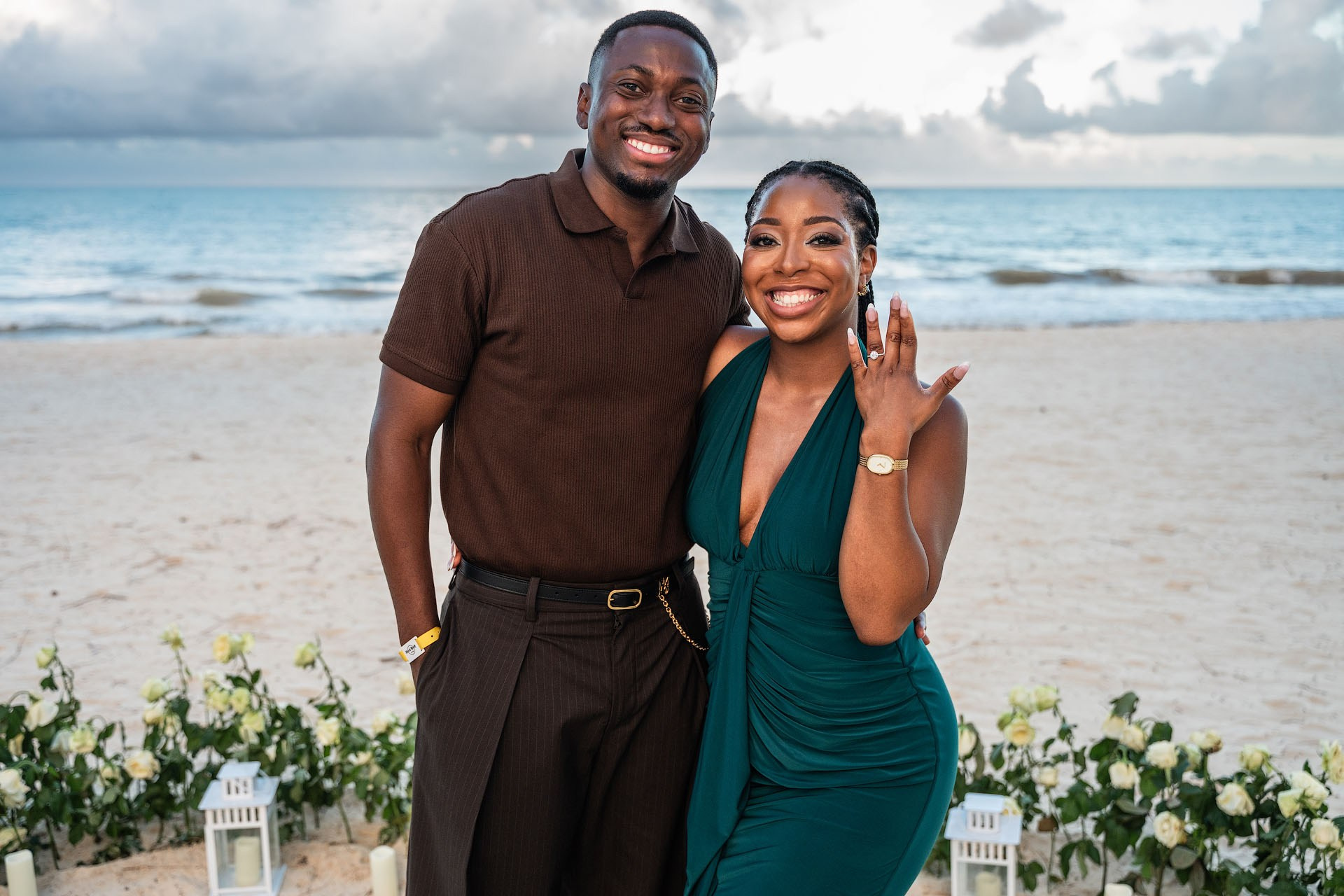 Smiling engaged couple posing by the ocean as one partner shows her engagement ring.
