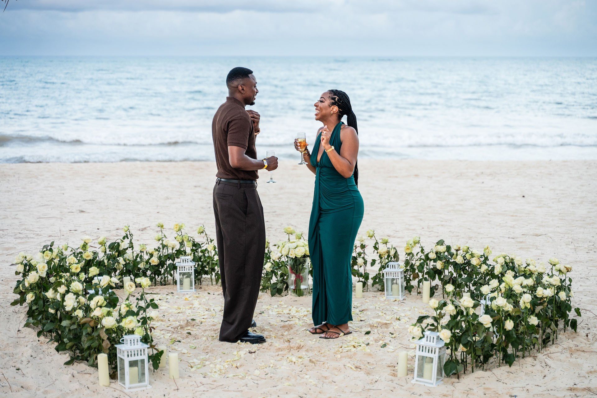 Newly engaged couple celebrating with champagne inside a heart-shaped arrangement of white roses on the beach.