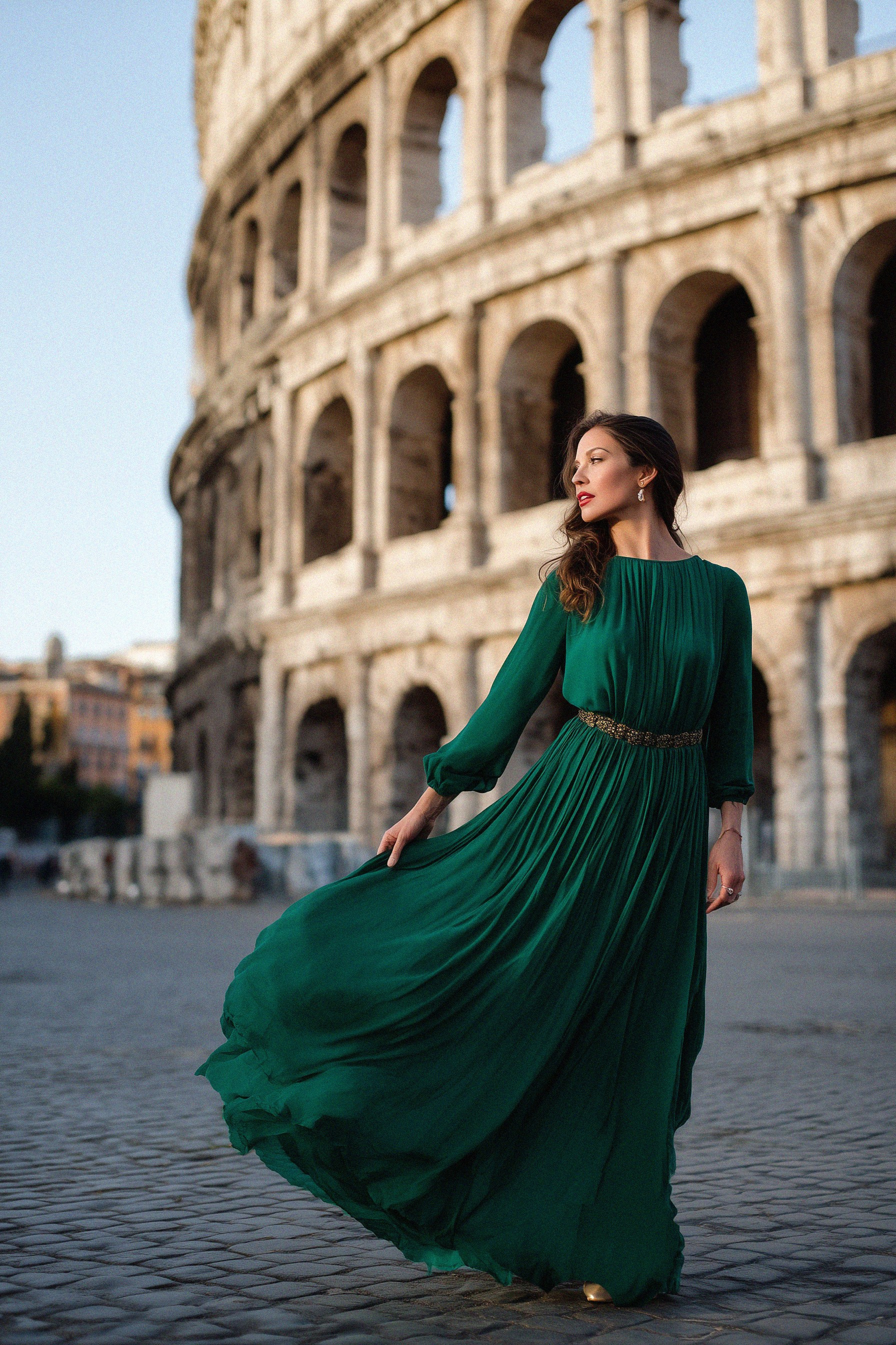 A woman in a flowing emerald green dress poses gracefully in front of the Colosseum in Rome during golden hour.