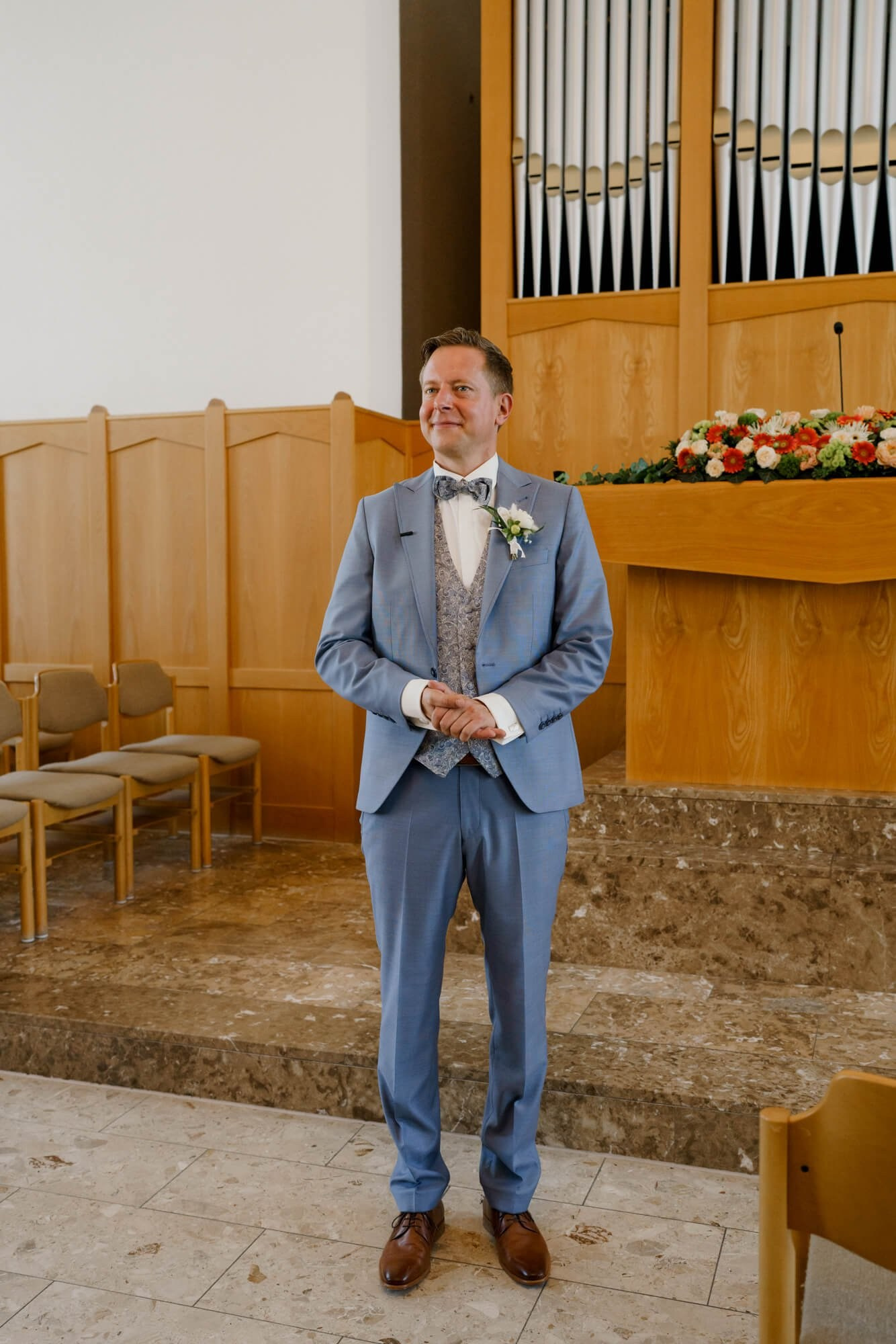 Groom standing at the front of the ceremony hall in a blue suit, a flower arrangement and organ pipes behind him