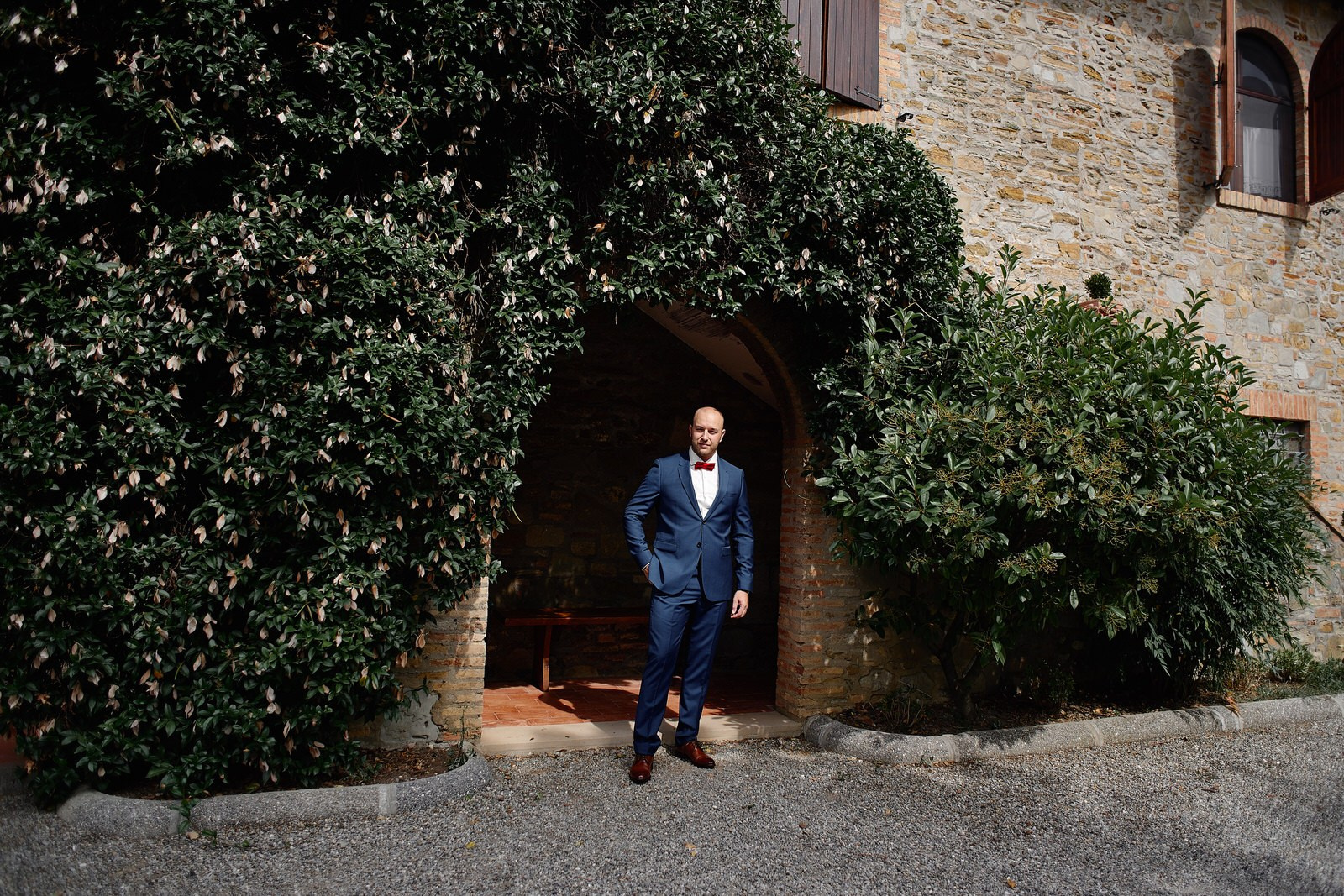 Groom in a navy blue suit standing at the ivy-covered arched entrance of Villa Barbara agriturismo, Tuscany, on his wedding morning