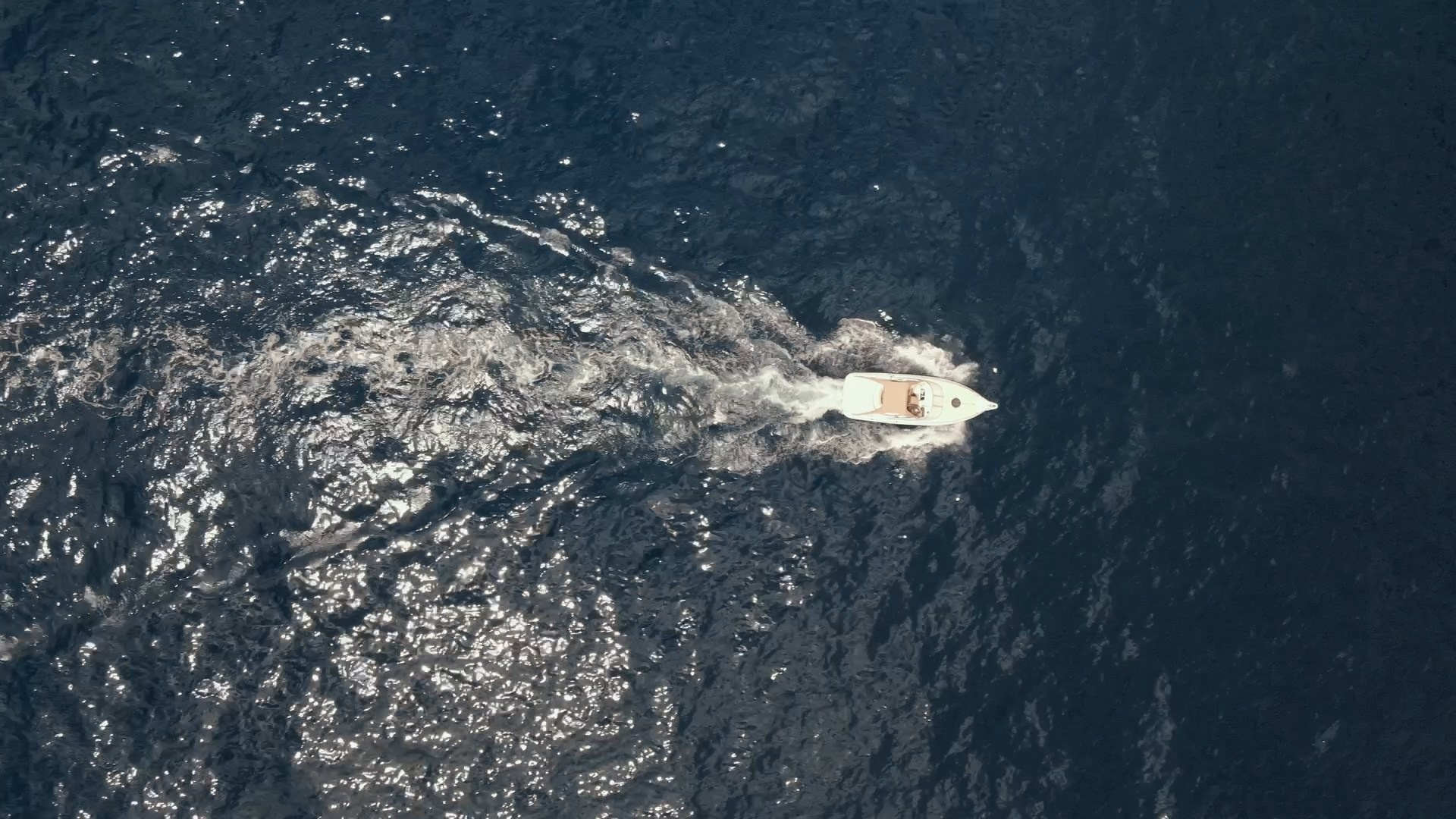 A top-down aerial view of a small white boat cutting through dark blue ocean waters, leaving a trail of gentle waves.