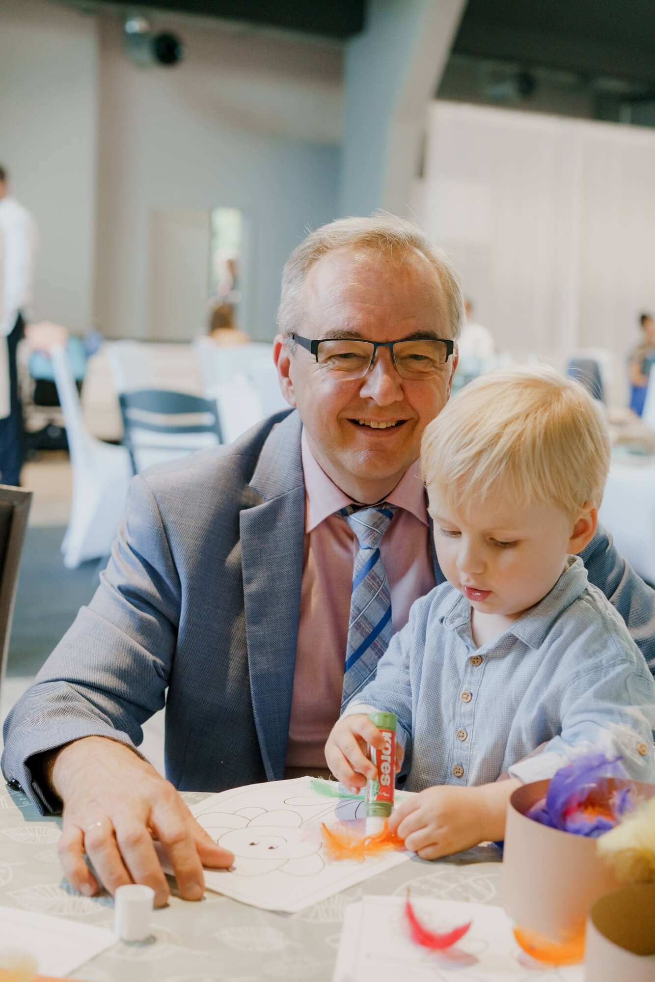 A wedding guest in glasses sitting with a young child who is drawing at a table during the reception