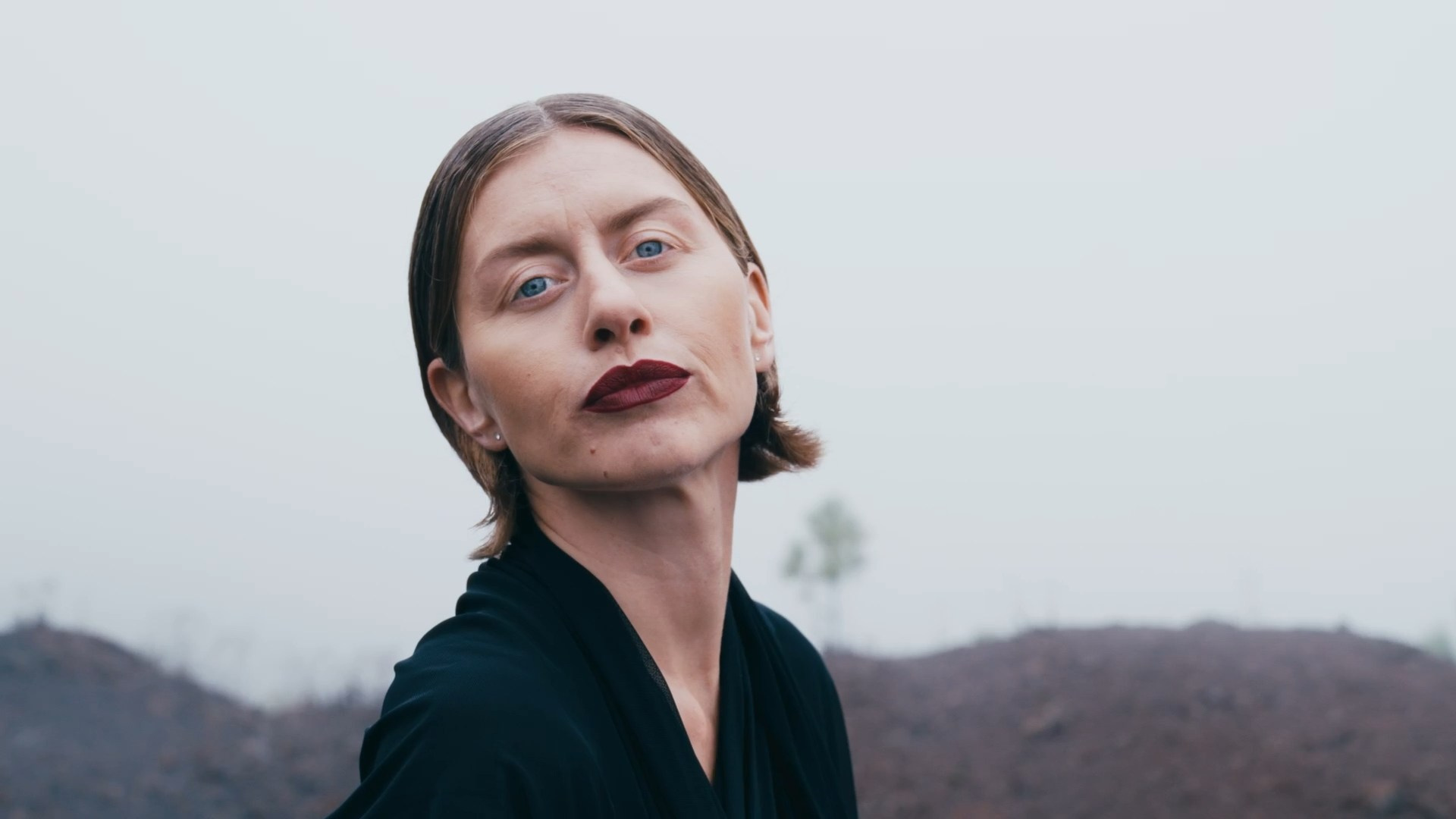 A close-up portrait of a woman with slicked-back hair and bold dark lipstick, set against a foggy outdoor background, exuding an elegant and introspective mood.