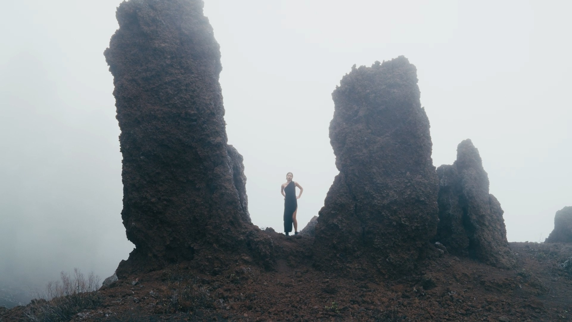 A misty landscape with towering volcanic rock formations. A lone figure dressed in black stands between the formations, creating a dramatic and minimalist composition.
