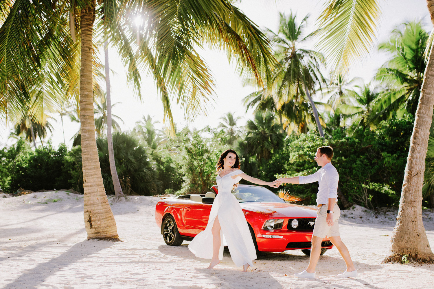 Невеста ведет жениха за руку в дольщика пляжа, за парой стоит красный мустанг специально арендованный для свадебной церемонии. 