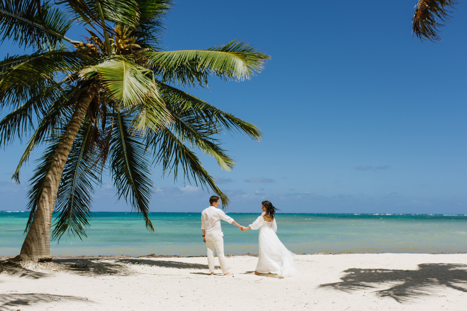 Пара позирует фотографу взявшись за руки. У невесты белое платье а у жениха.