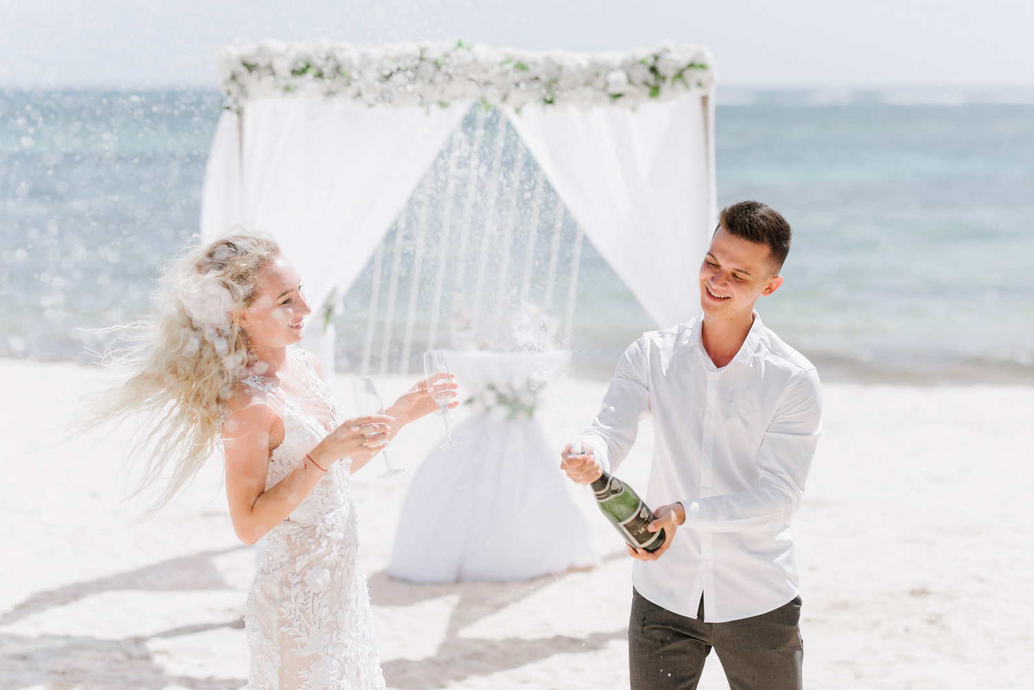 Невеста и жених открывают бутылку шампанского на пляже.