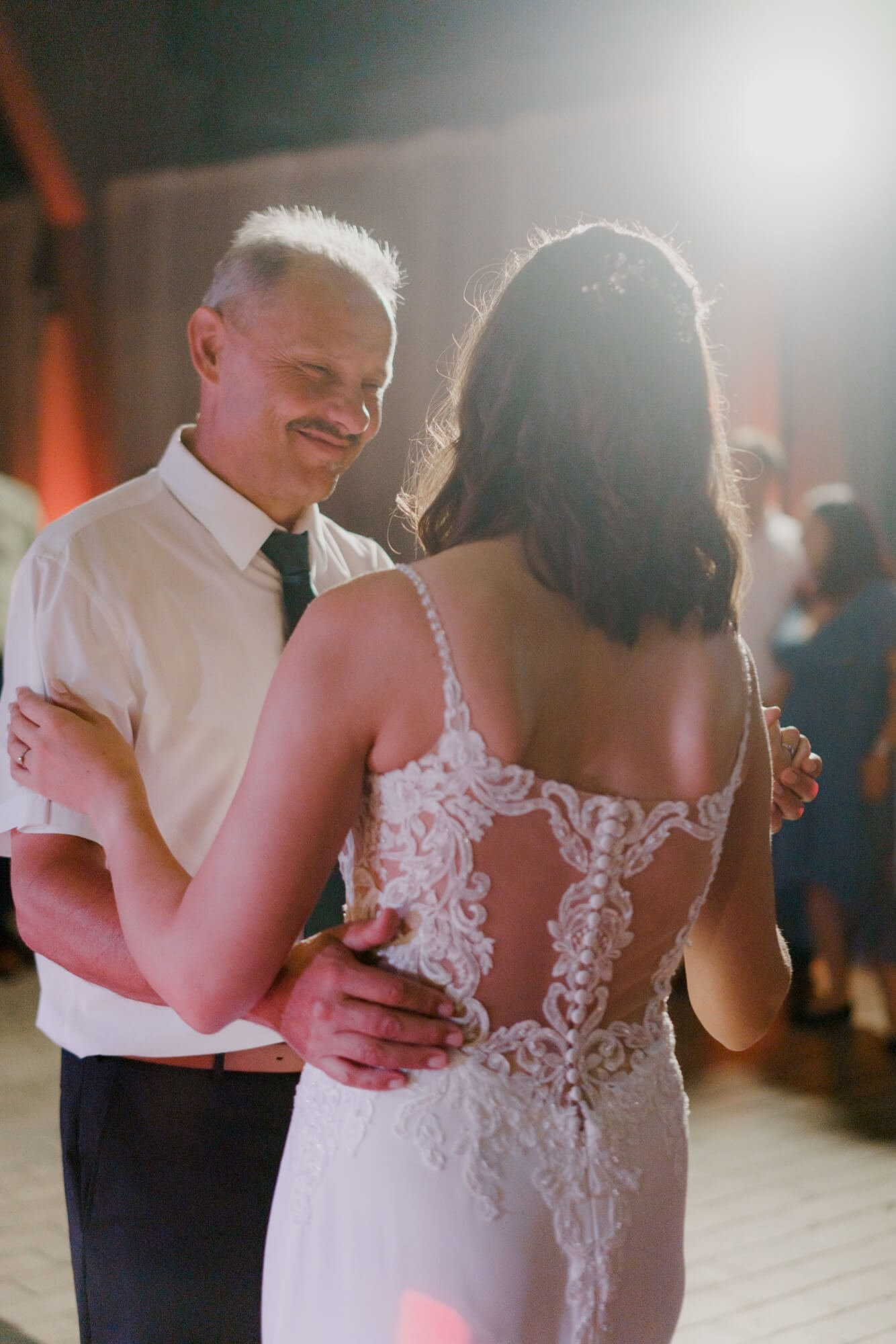 Bride dancing with an older male guest, the intricate lace back of her gown prominent in the warm reception light