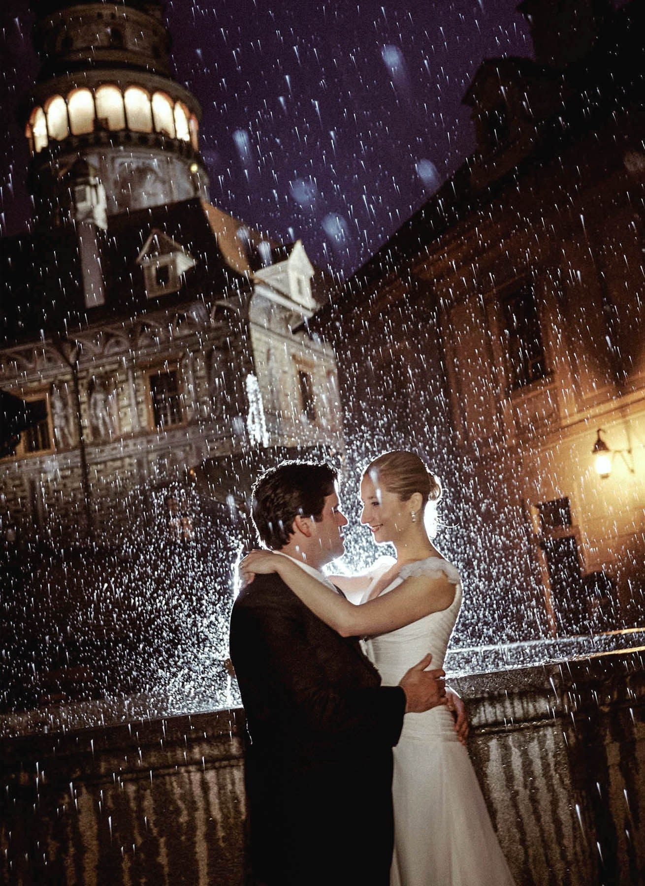 Gorgeous newlyweds share a tender embrace in the rain within the historic courtyard of Cesky Krumlov Castle, Czechia, on their wedding day.