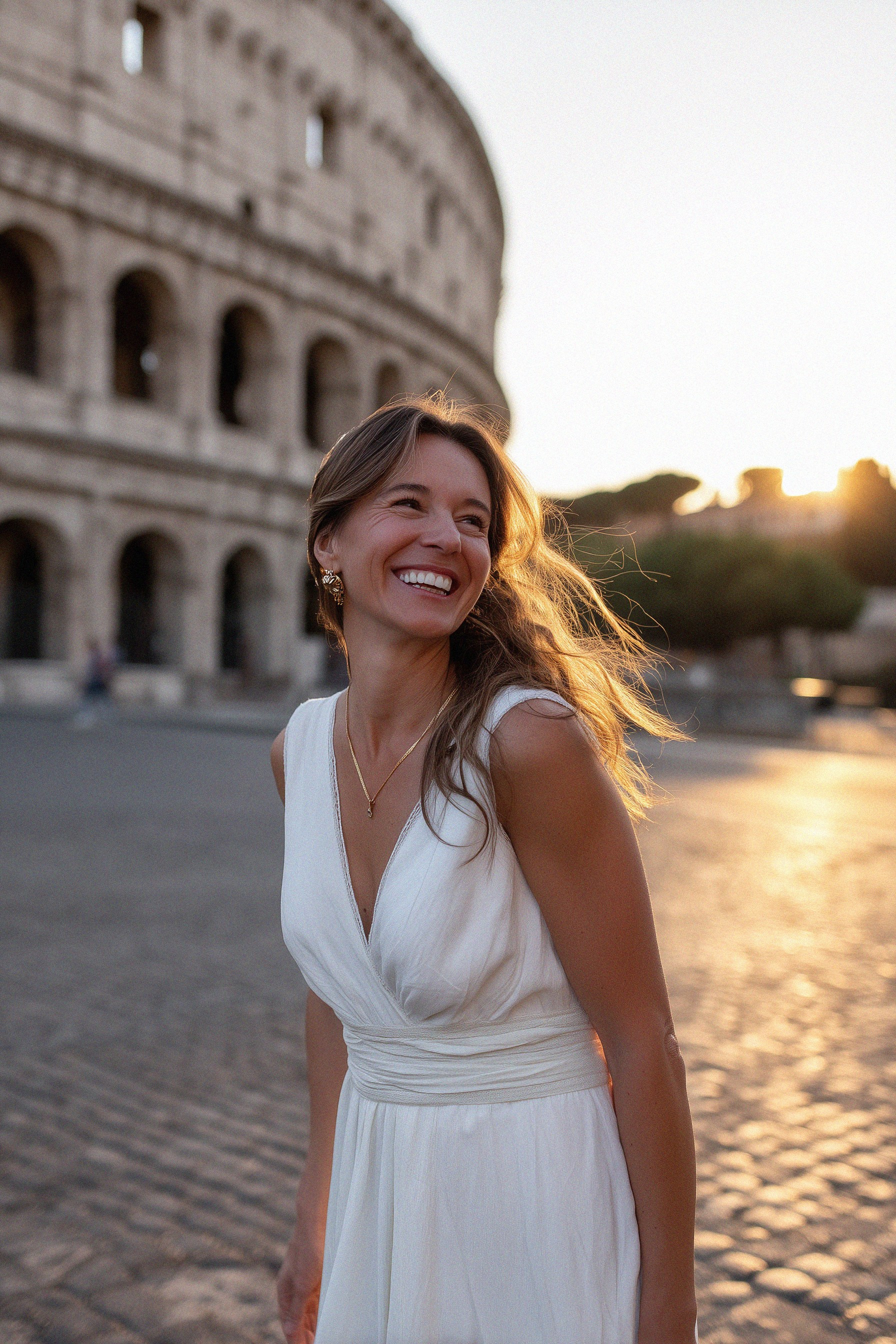 A smiling woman in a white dress stands near the Colosseum at sunrise, bathed in golden light.