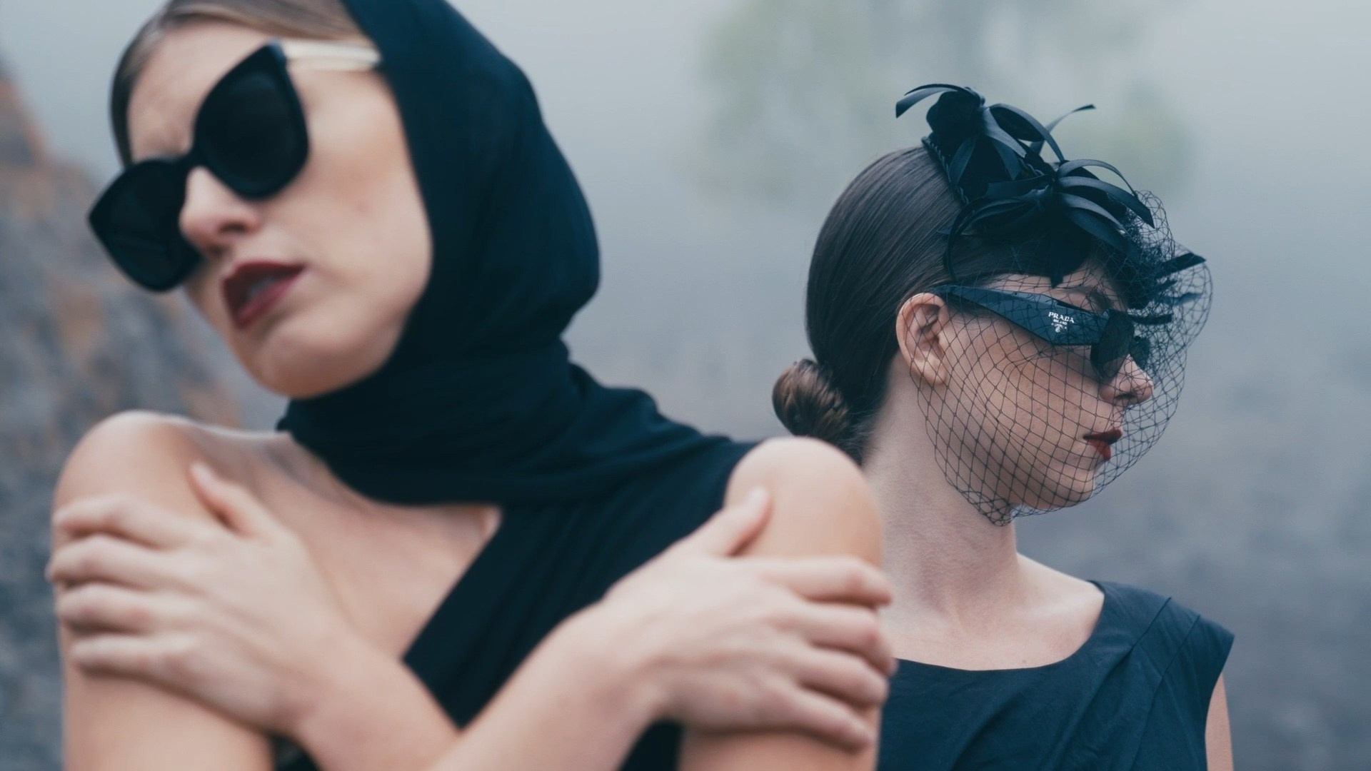 Two women in dark, dramatic attire. The foreground features a woman wearing oversized black sunglasses and a scarf, embracing herself, while the background shows another woman with a decorative headpiece and veil, adding a mysterious and artistic tone.