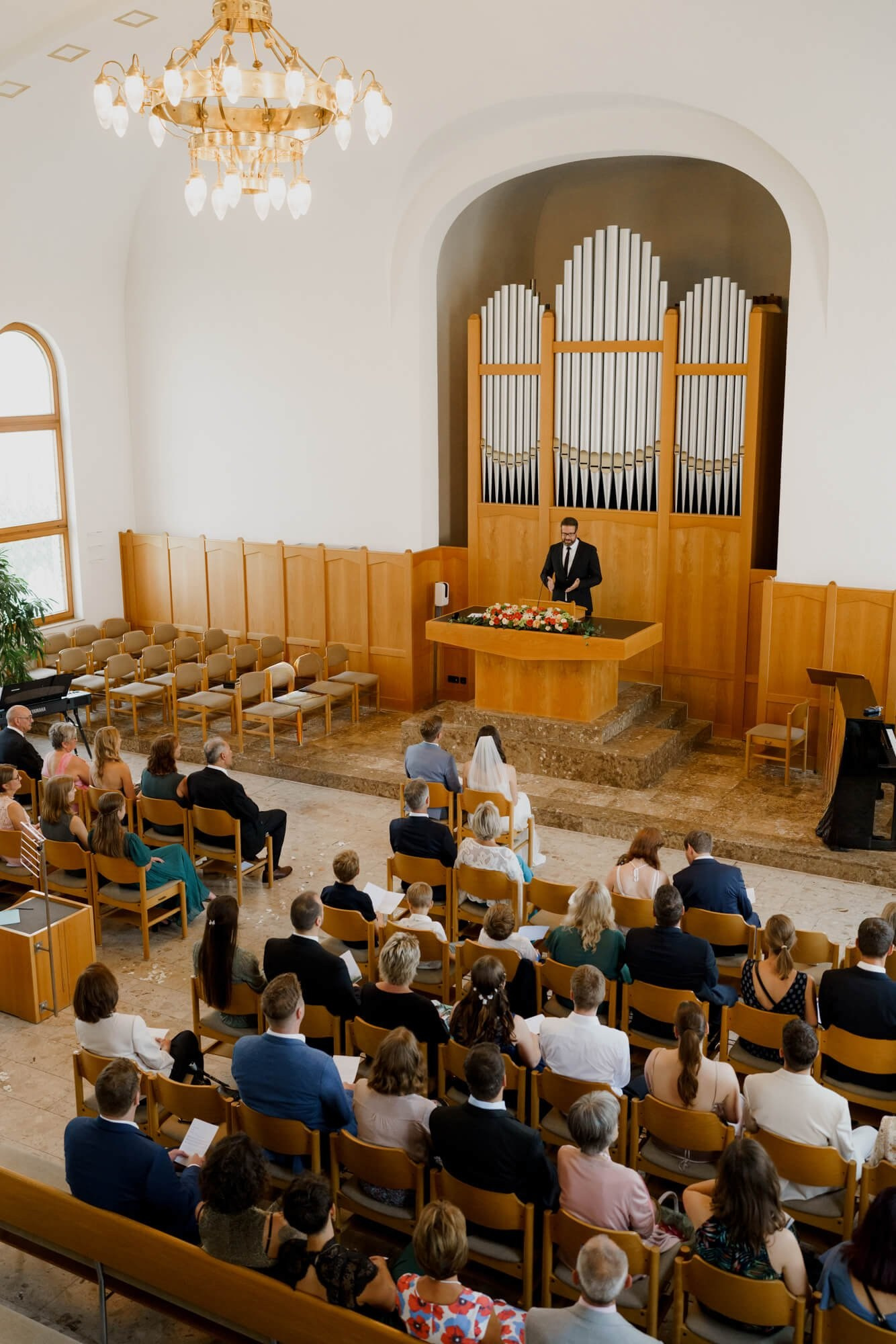 Overhead view of the full Stuttgart ceremony hall showing guests seated in wooden pews, a pipe organ at the rear, and a chandelier overhead