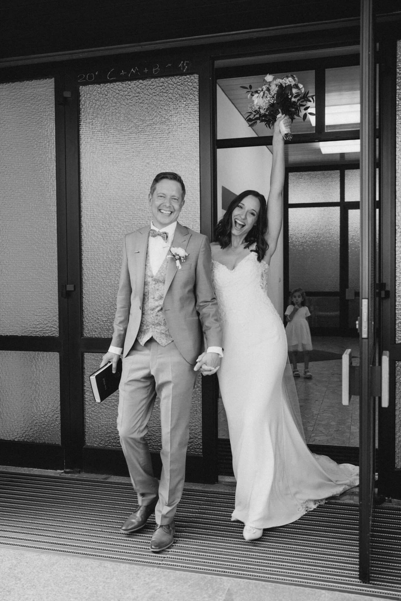 Bride raising her bouquet high as the couple bursts through the ceremony exit doors together, both laughing; black and white