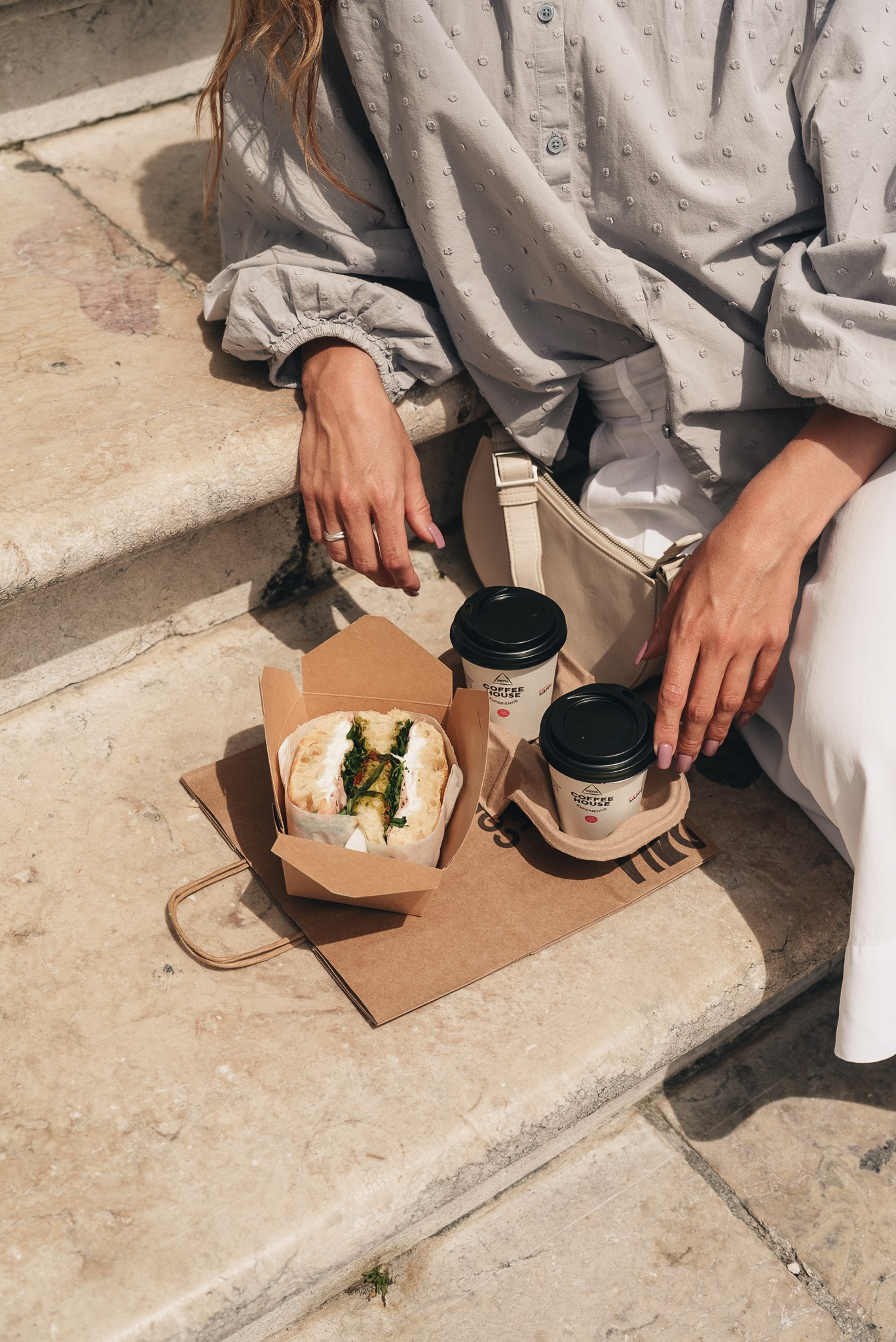 Fotografia lifestyle urbana de uma pessoa sentada em escadas de pedra com um lanche de take-away e copos de café, pela Khave Media
