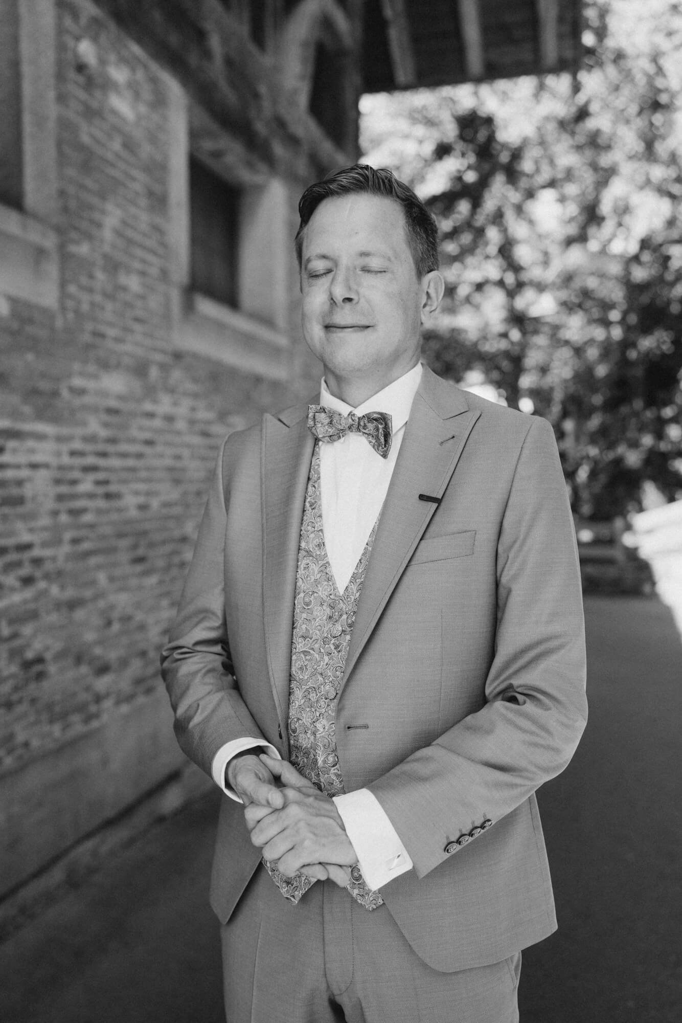 Groom standing alone with eyes closed and hands clasped outside a historic brick building; black and white