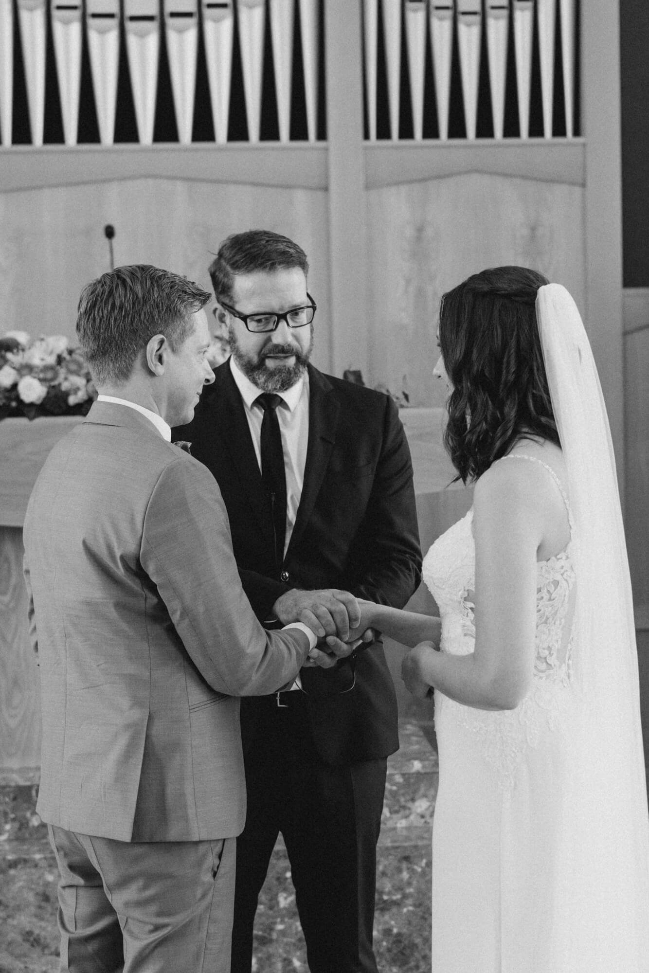 Couple holding hands at the altar as an officiant in glasses reads during the Stuttgart wedding ceremony; black and white