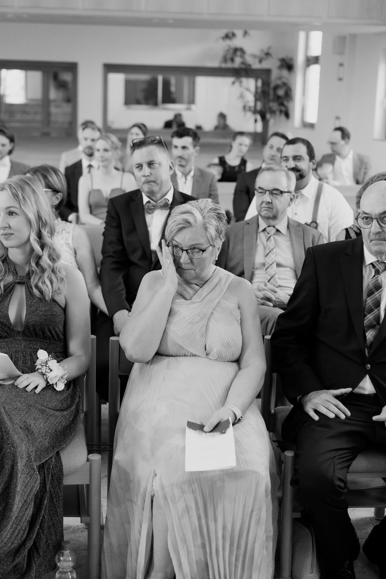 Wedding guests seated in the ceremony hall, a woman in the foreground wiping her eye during the vows; black and white